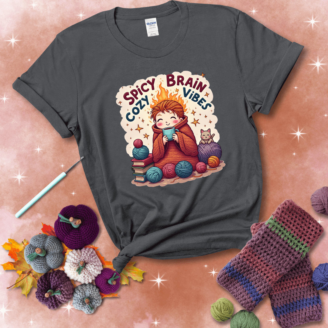 A person with flaming red hair wrapped in a chunky crochet blanket, holding a mug, surrounded by yarn balls, books, a cat, and the phrase "Spicy Brain Cozy Vibes" showing a dark grey tee with a festive Fall background
