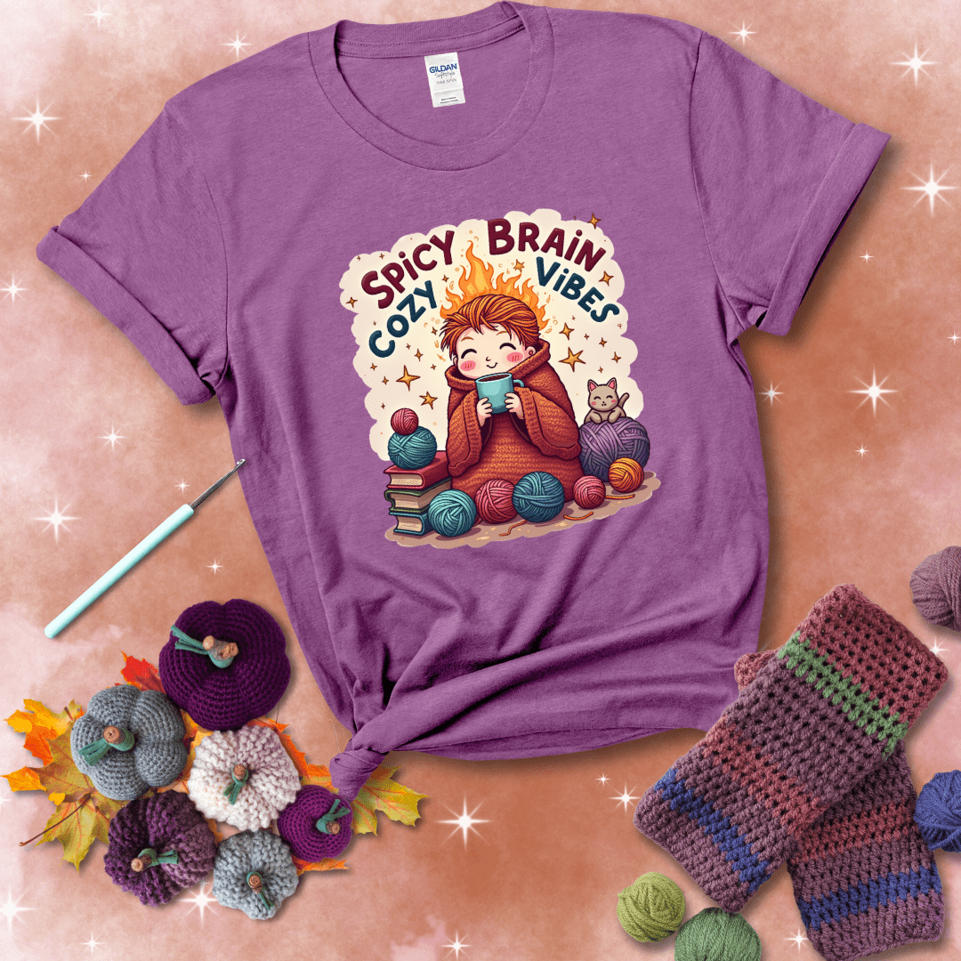A person with flaming red hair wrapped in a chunky crochet blanket, holding a mug, surrounded by yarn balls, books, a cat, and the phrase "Spicy Brain Cozy Vibes" showing a light purple tee with a festive Fall background