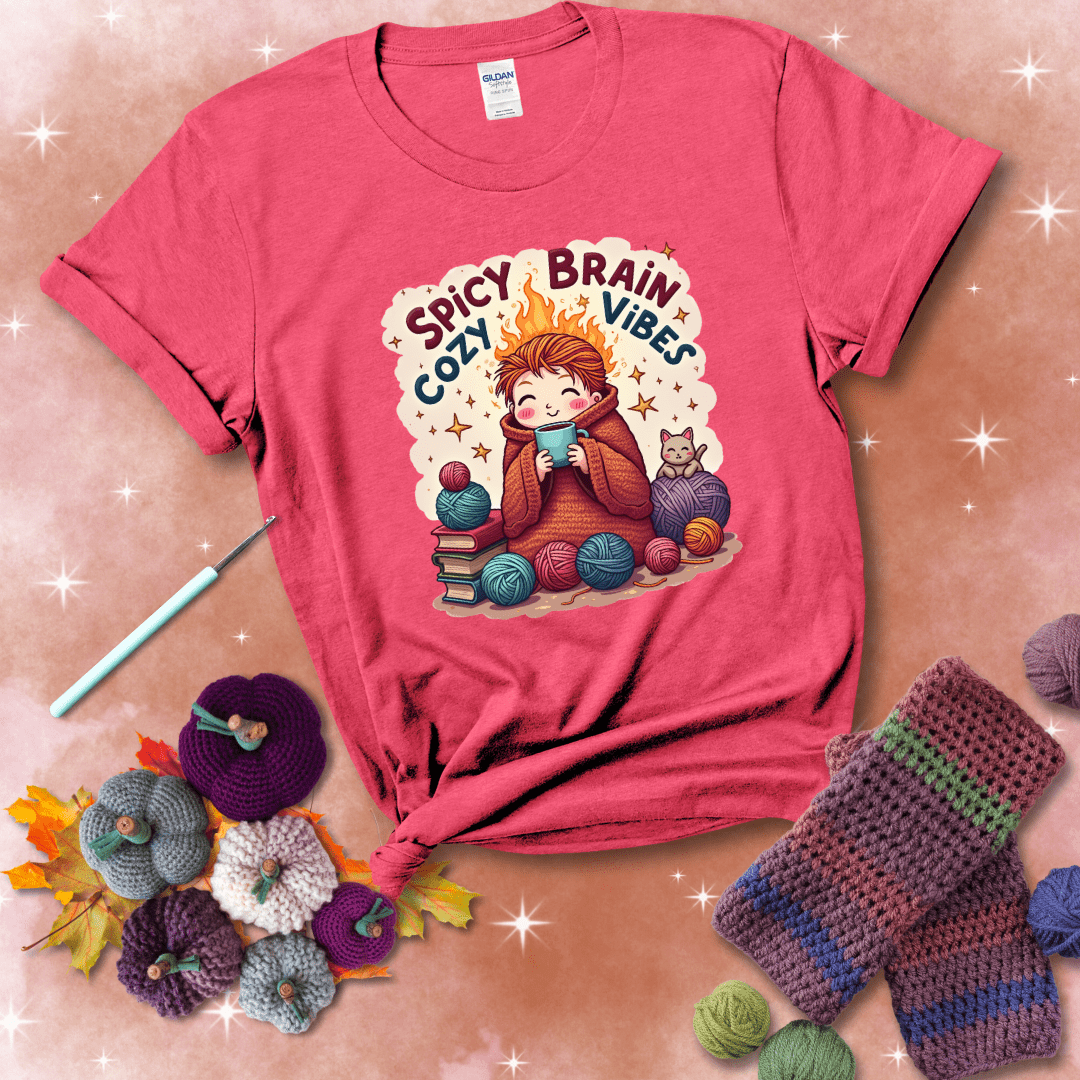 A person with flaming red hair wrapped in a chunky crochet blanket, holding a mug, surrounded by yarn balls, books, a cat, and the phrase "Spicy Brain Cozy Vibes" showing a light red tee with a festive Fall background