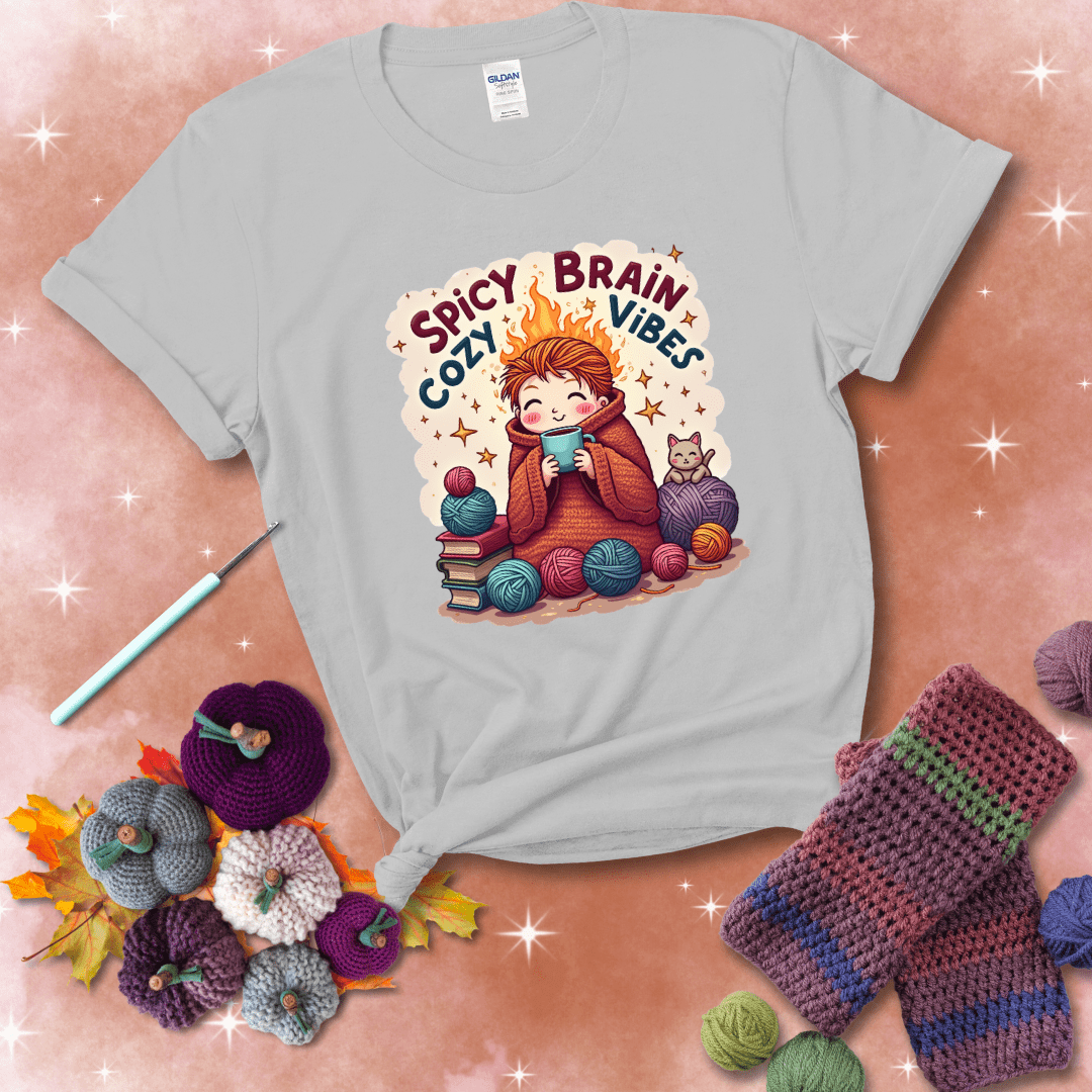 A person with flaming red hair wrapped in a chunky crochet blanket, holding a mug, surrounded by yarn balls, books, a cat, and the phrase "Spicy Brain Cozy Vibes" showing a light grey tee with a festive Fall background