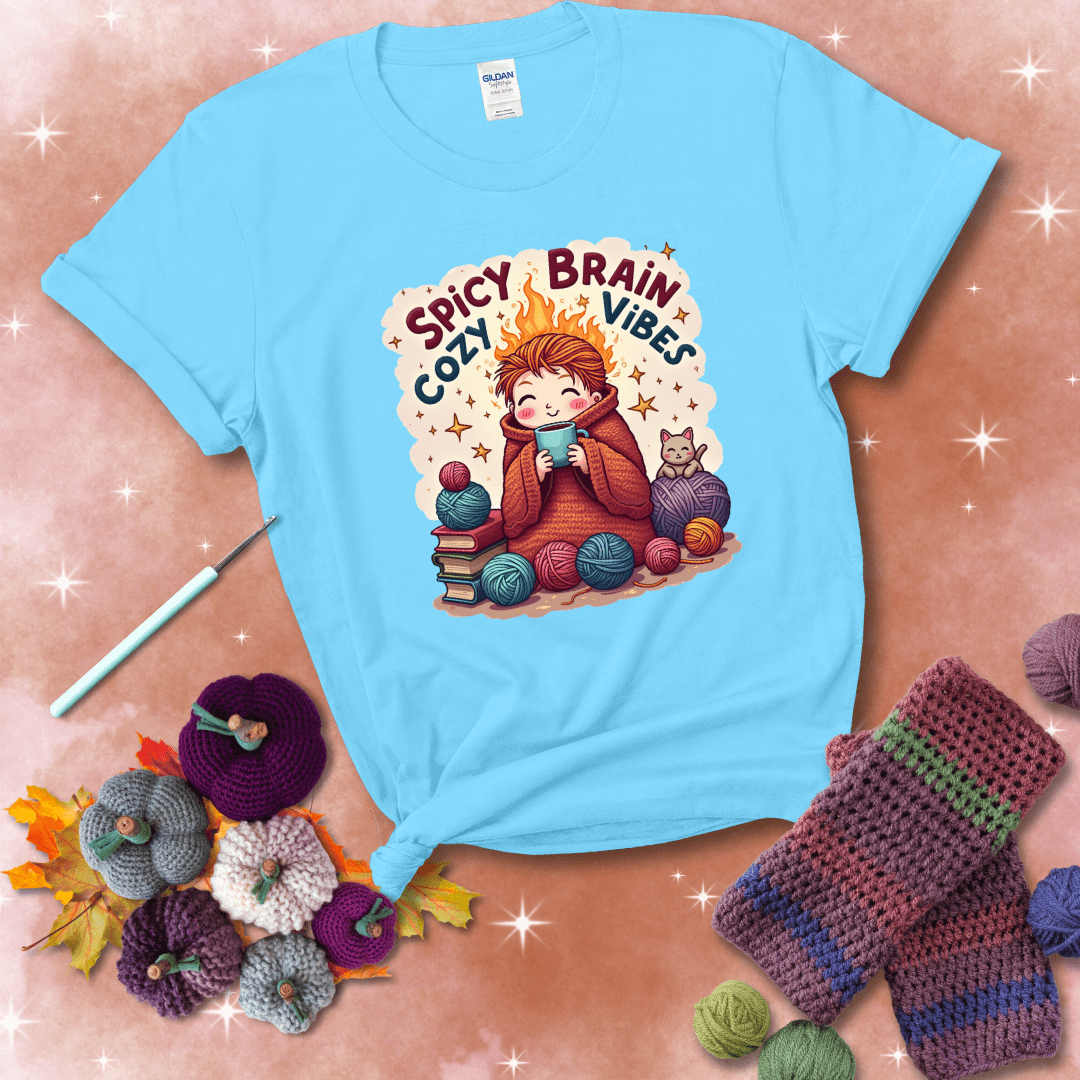 A person with flaming red hair wrapped in a chunky crochet blanket, holding a mug, surrounded by yarn balls, books, a cat, and the phrase "Spicy Brain Cozy Vibes" showing a light blue tee with a festive Fall background