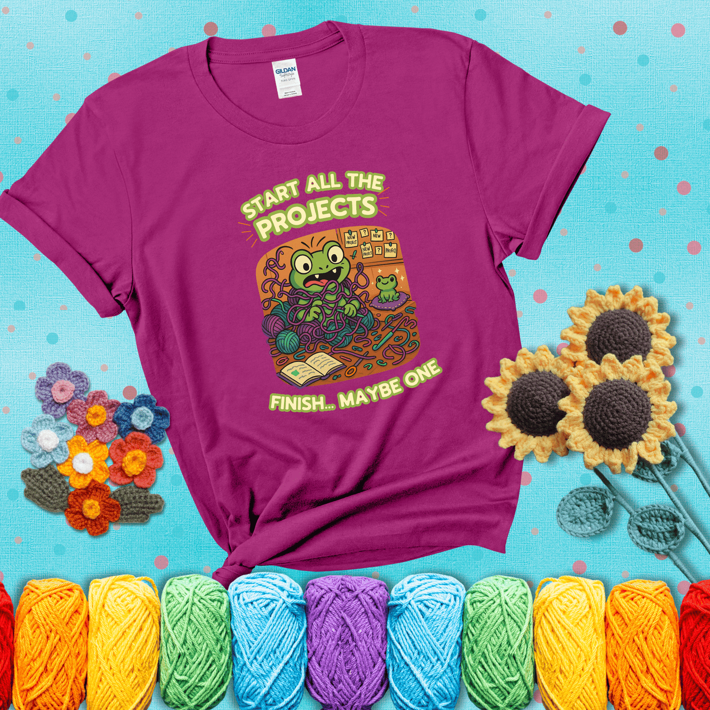 A wide-eyed crochet frog sits tangled in yarn with dozens of projects around, surrounded by chaos and “new project” notes. Caption reads “Start all the projects, finish... maybe one” showing a magenta tee with a teal sunflower background