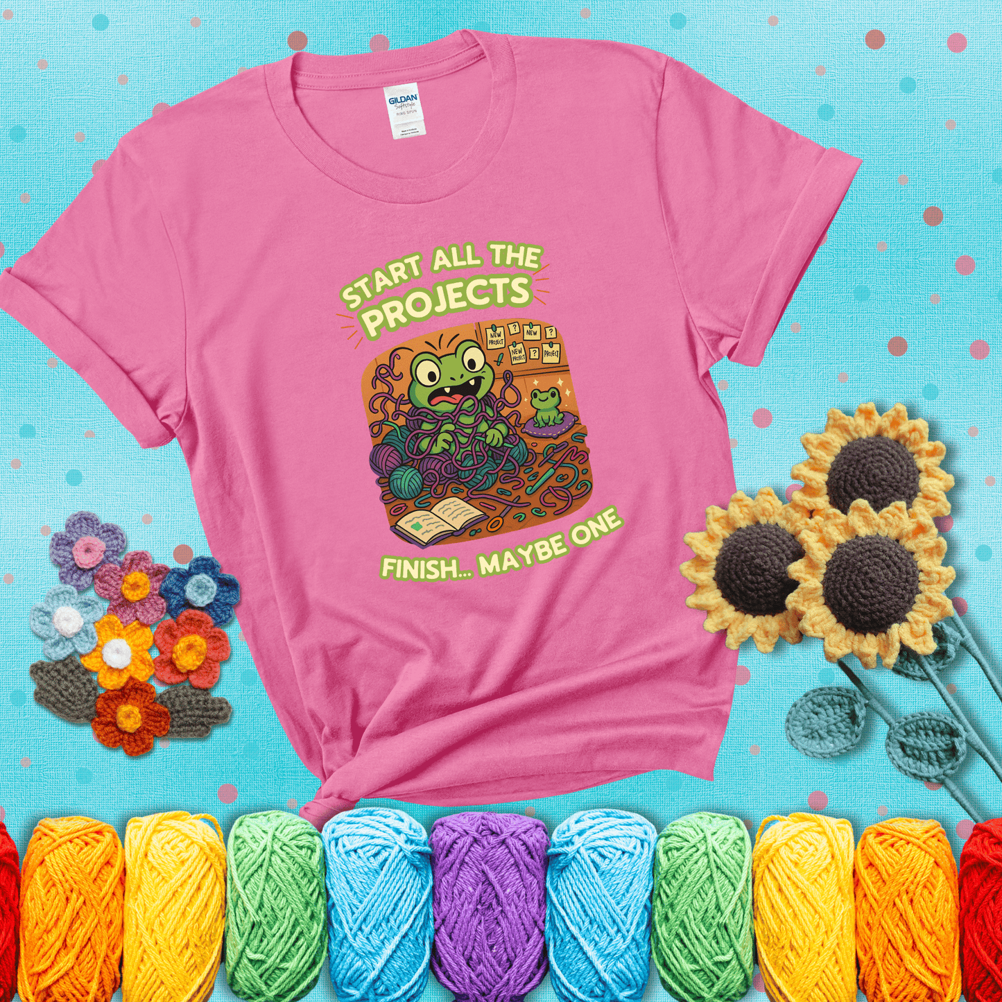 A wide-eyed crochet frog sits tangled in yarn with dozens of projects around, surrounded by chaos and “new project” notes. Caption reads “Start all the projects, finish... maybe one” showing a pink tee with a teal sunflower background