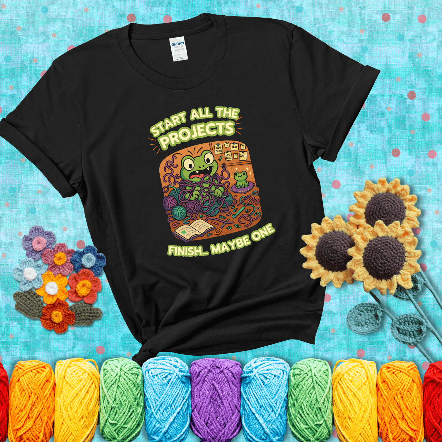 A wide-eyed crochet frog sits tangled in yarn with dozens of projects around, surrounded by chaos and “new project” notes. Caption reads “Start all the projects, finish... maybe one” showing a black tee with a teal sunflower background