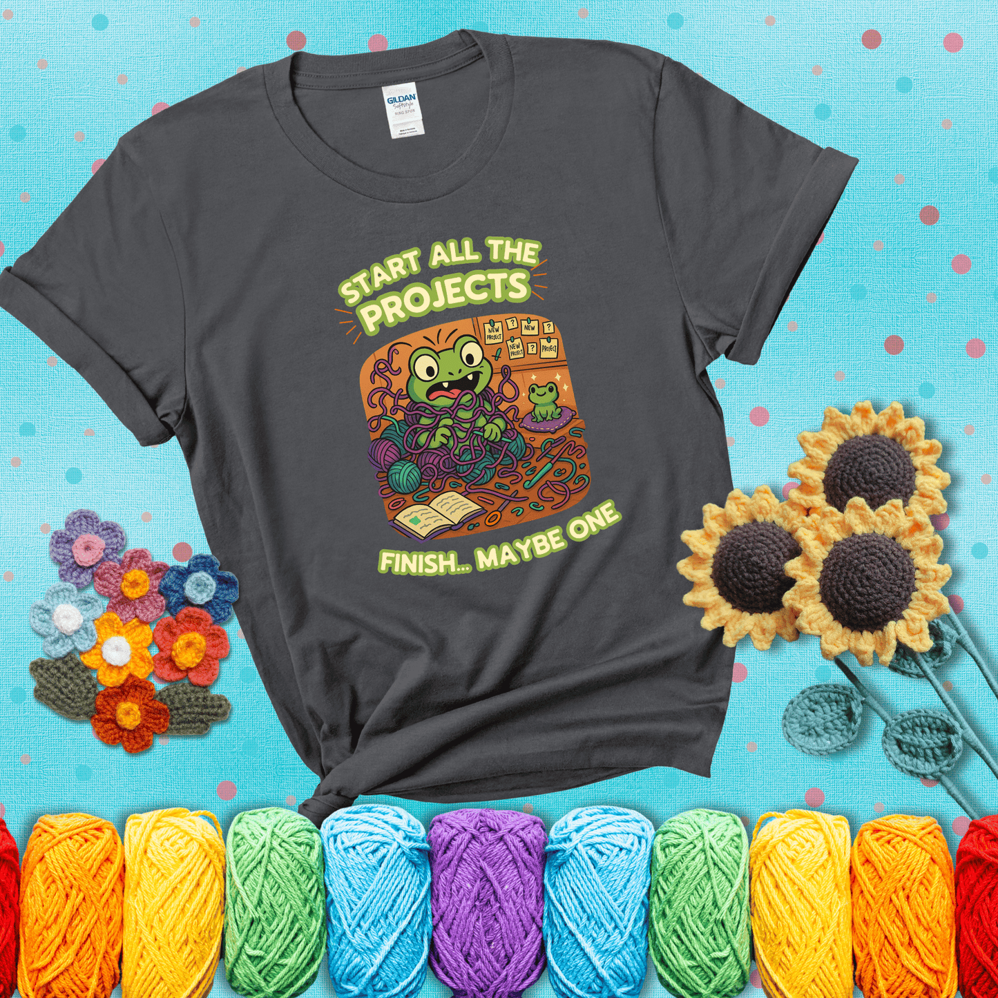 A wide-eyed crochet frog sits tangled in yarn with dozens of projects around, surrounded by chaos and “new project” notes. Caption reads “Start all the projects, finish... maybe one” showing a dark grey tee with a teal sunflower background