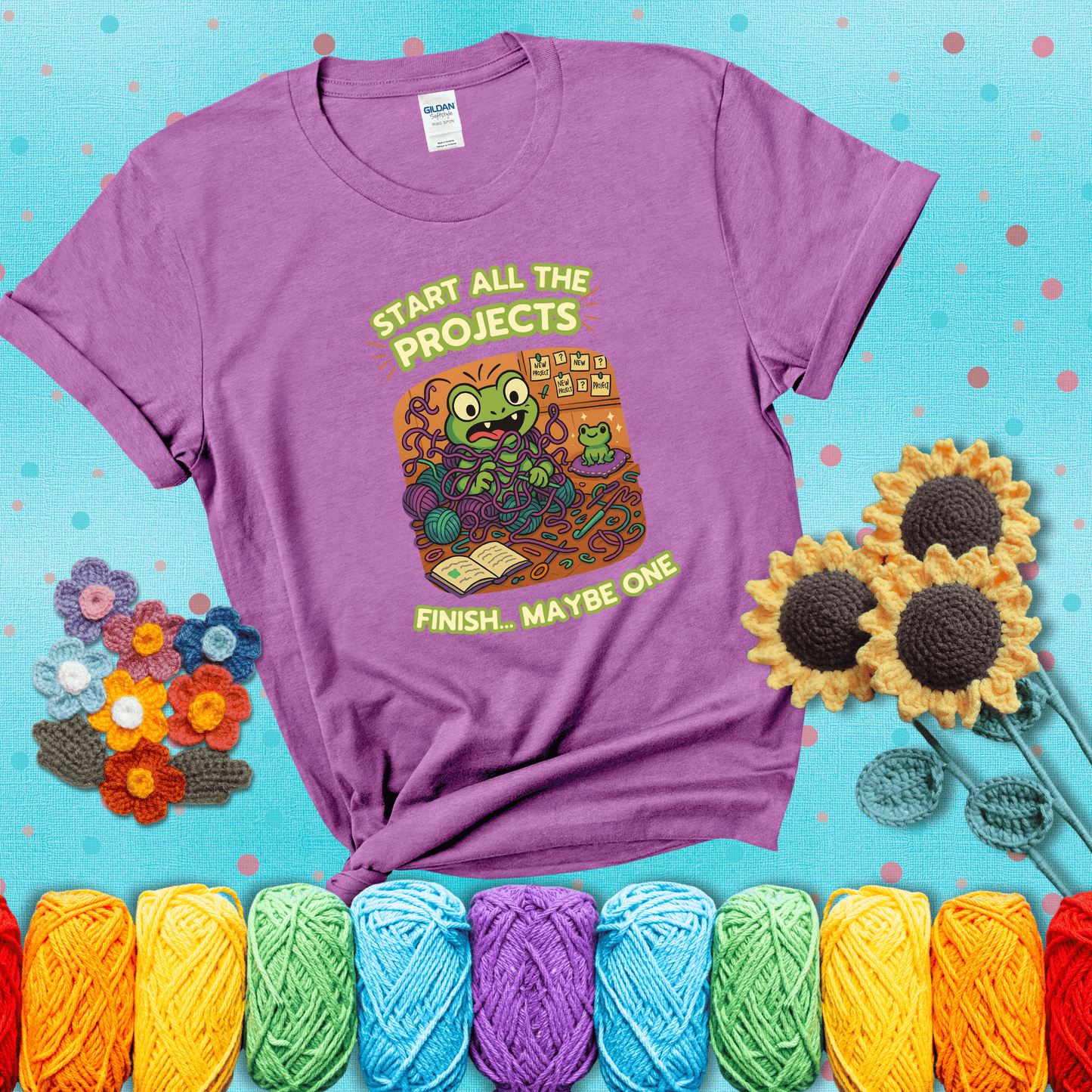 A wide-eyed crochet frog sits tangled in yarn with dozens of projects around, surrounded by chaos and “new project” notes. Caption reads “Start all the projects, finish... maybe one” showing a light purple tee with a teal sunflower background