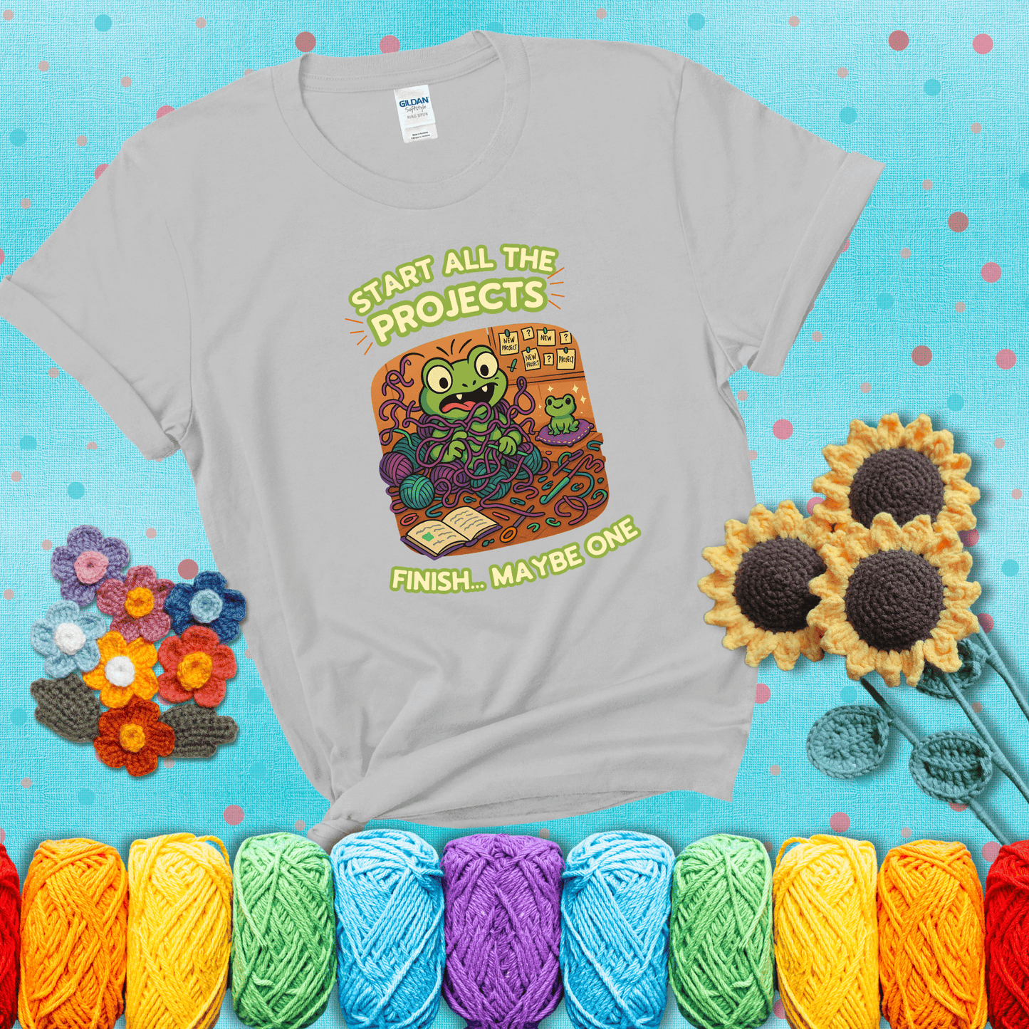 A wide-eyed crochet frog sits tangled in yarn with dozens of projects around, surrounded by chaos and “new project” notes. Caption reads “Start all the projects, finish... maybe one” showing a light grey tee with a teal sunflower background