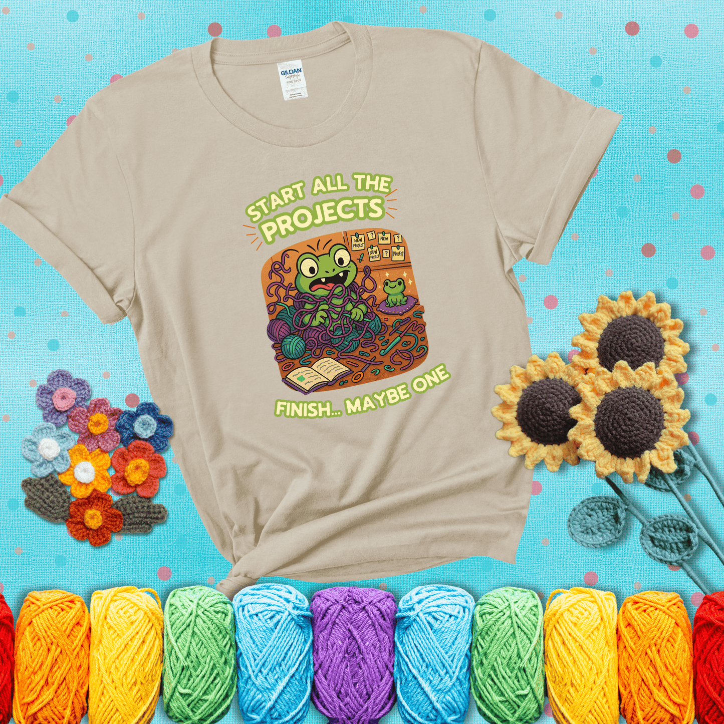 A wide-eyed crochet frog sits tangled in yarn with dozens of projects around, surrounded by chaos and “new project” notes. Caption reads “Start all the projects, finish... maybe one” showing a beige tee with a teal sunflower background