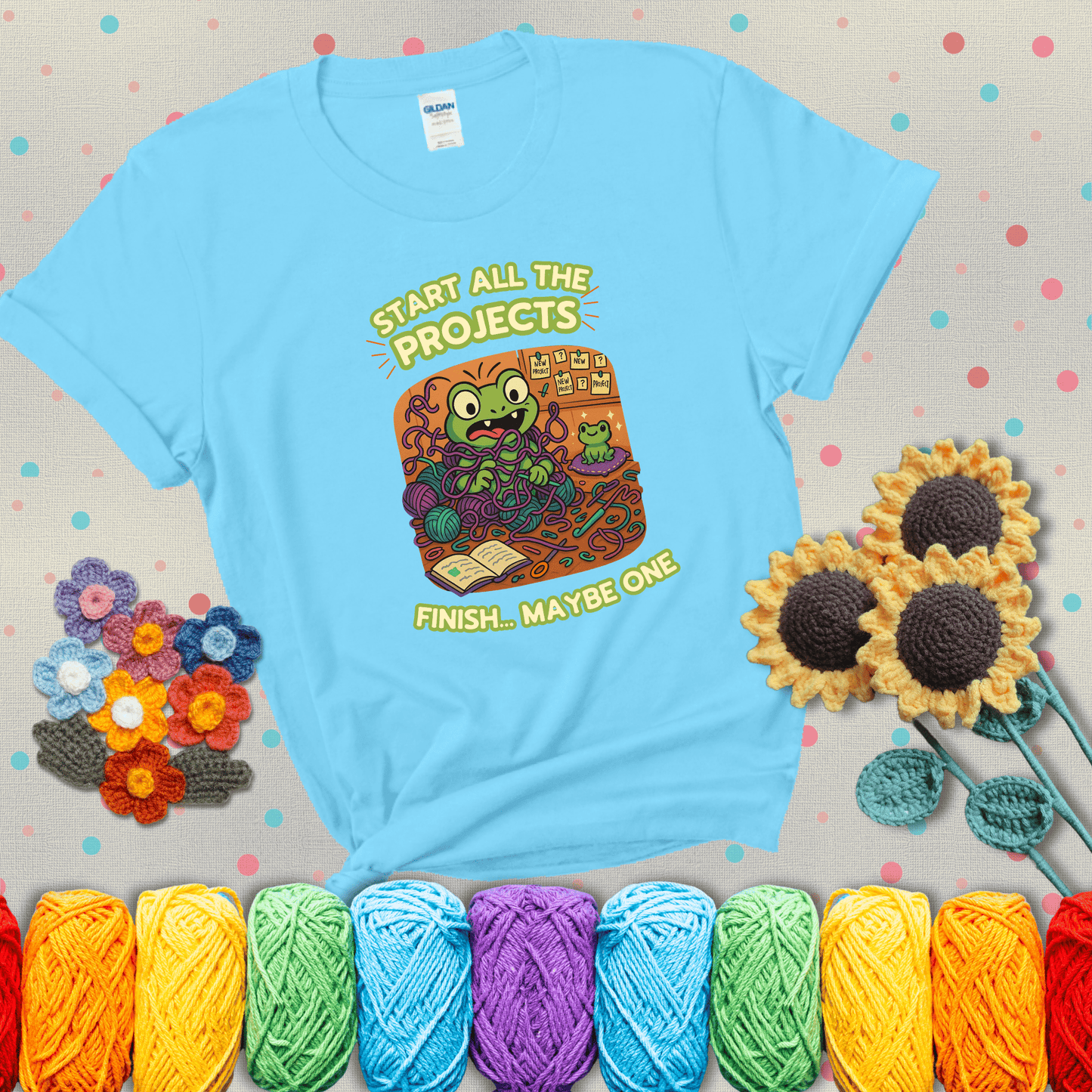 A wide-eyed crochet frog sits tangled in yarn with dozens of projects around, surrounded by chaos and “new project” notes. Caption reads “Start all the projects, finish... maybe one” showing a light blue tee with grey sunflower background
