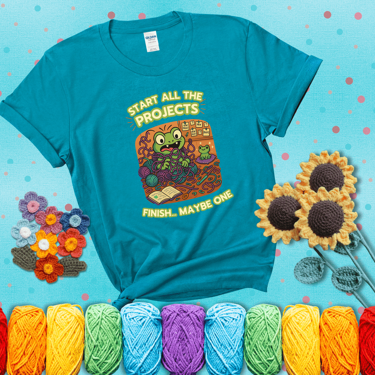 A wide-eyed crochet frog sits tangled in yarn with dozens of projects around, surrounded by chaos and “new project” notes. Caption reads “Start all the projects, finish... maybe one” showing a blue-green tee with a teal sunflower background