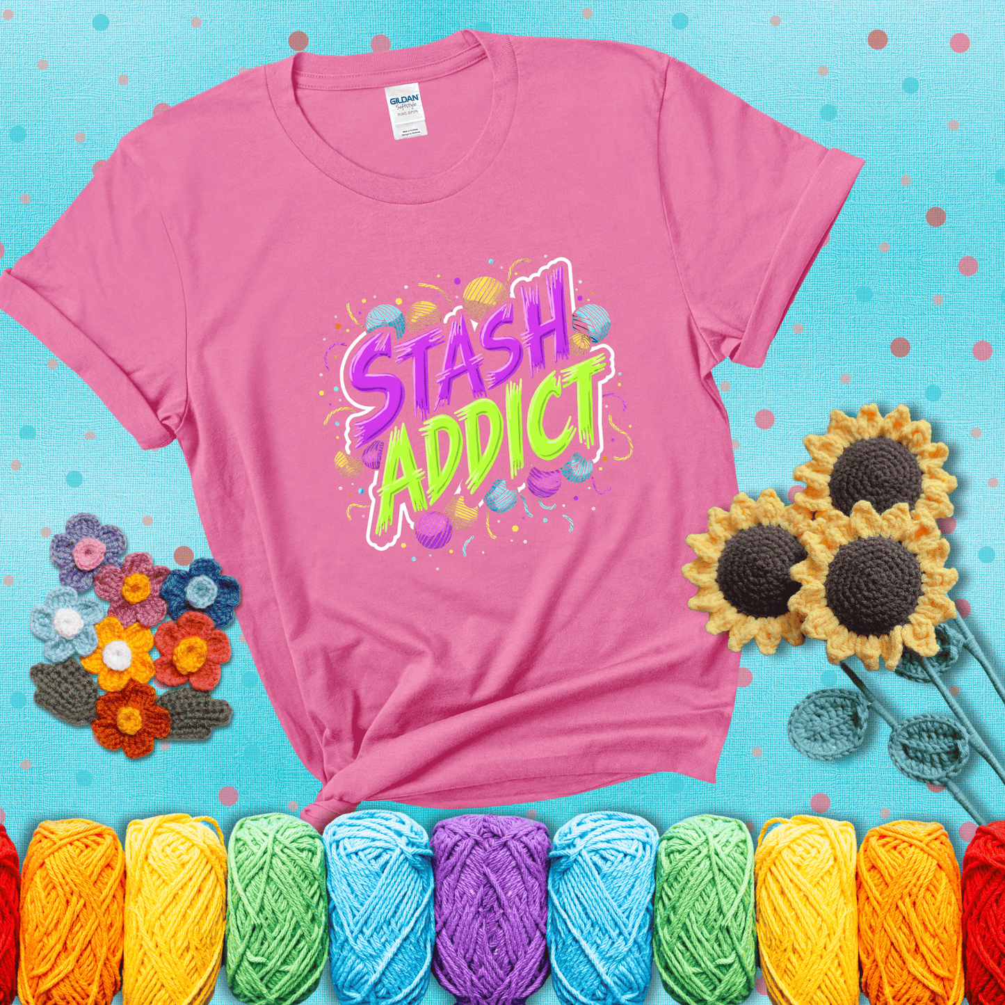 Bold neon text reading "Stash Addict" surrounded by colorful yarn balls and festive confetti on a neutral background showing a pink tee with a teal sunflower background