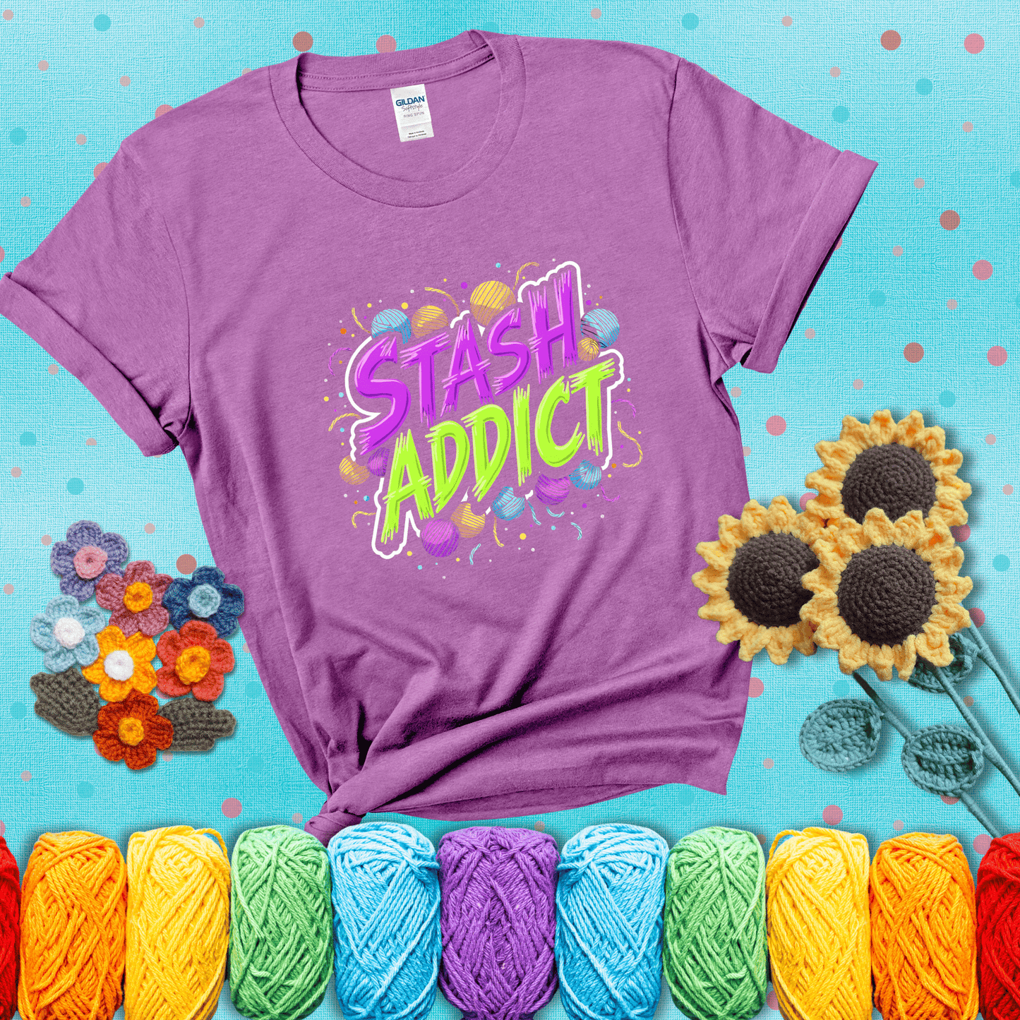 Bold neon text reading "Stash Addict" surrounded by colorful yarn balls and festive confetti on a neutral background showing a light purple tee with a teal sunflower background