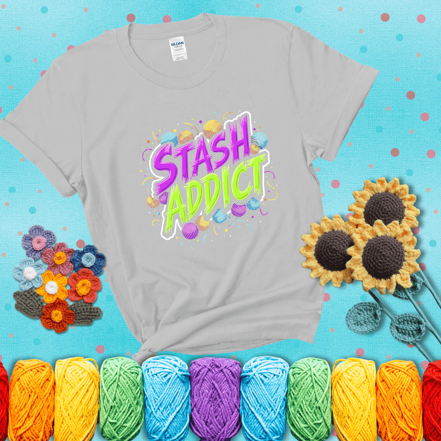 Bold neon text reading "Stash Addict" surrounded by colorful yarn balls and festive confetti on a neutral background showing a light grey tee with a teal sunflower background