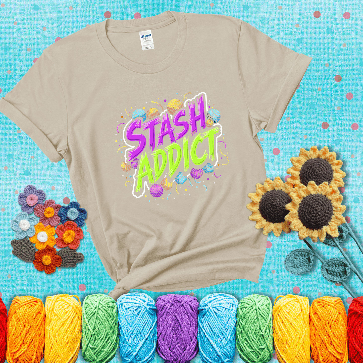 Bold neon text reading "Stash Addict" surrounded by colorful yarn balls and festive confetti on a neutral background showing a beige tee with a teal sunflower background