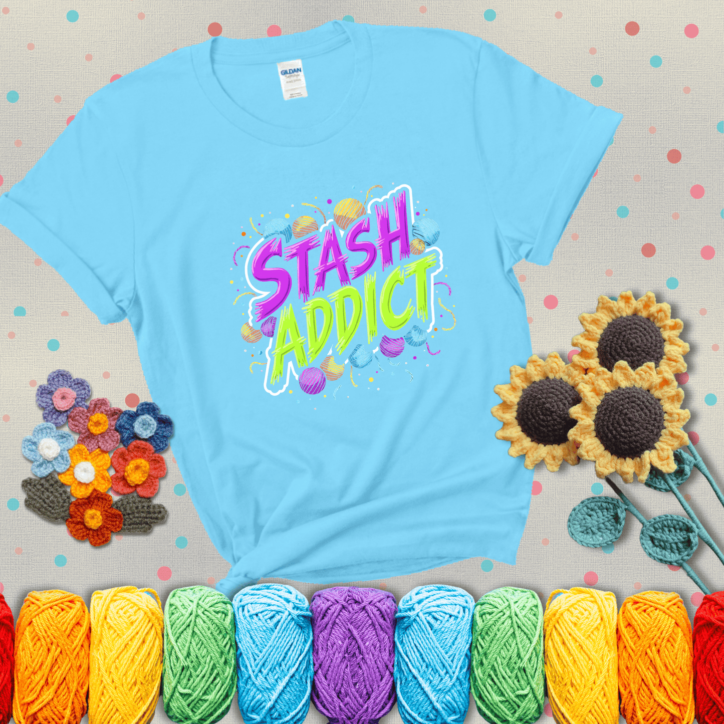 Bold neon text reading "Stash Addict" surrounded by colorful yarn balls and festive confetti on a neutral background showing a light blue tee with grey sunflower background