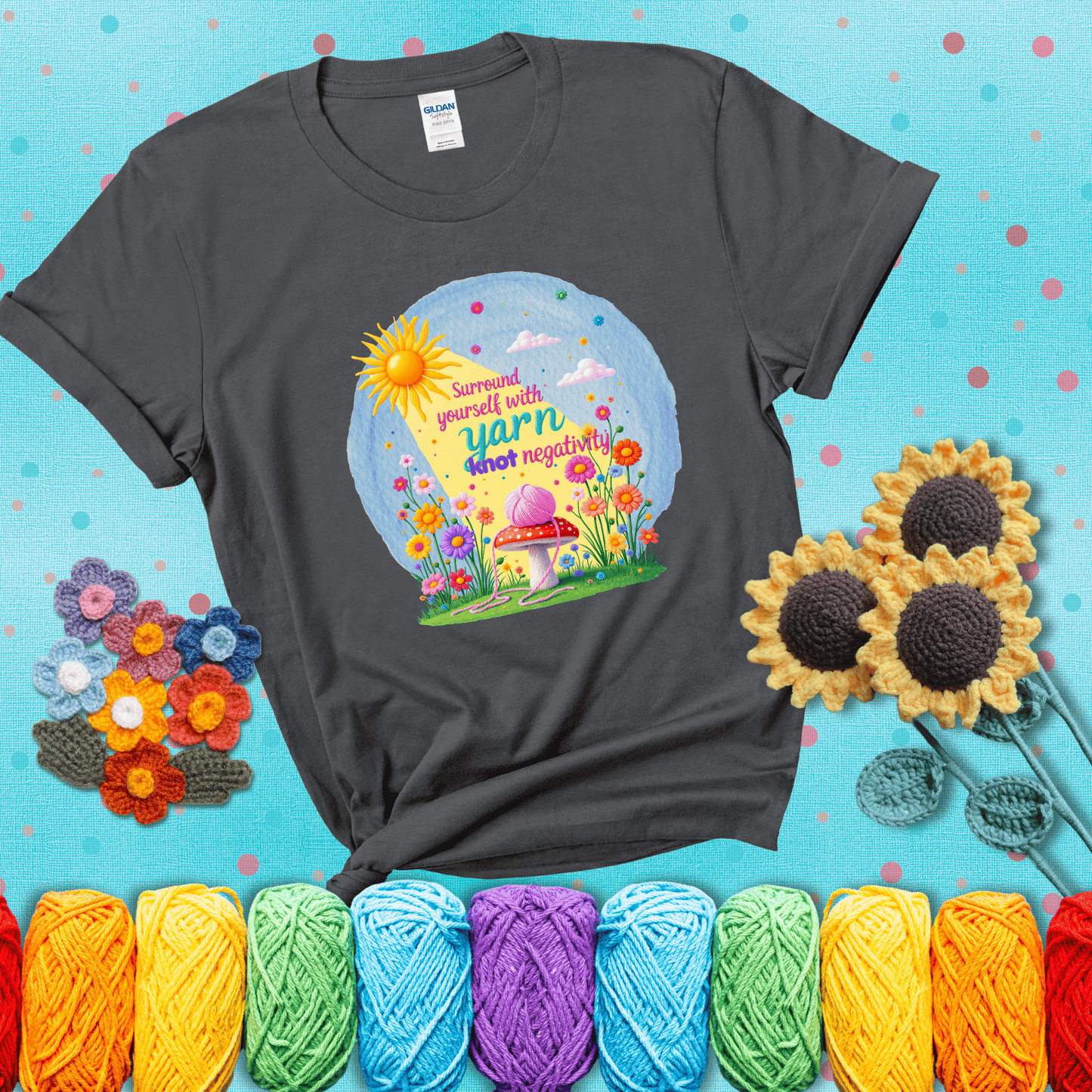 A happy spring garden scene with a sunbeam, flowers, and a pink yarn ball on a mushroom, with the quote "Surround yourself with yarn, knot negativity," showing a dark grey tee with a teal sunflower background