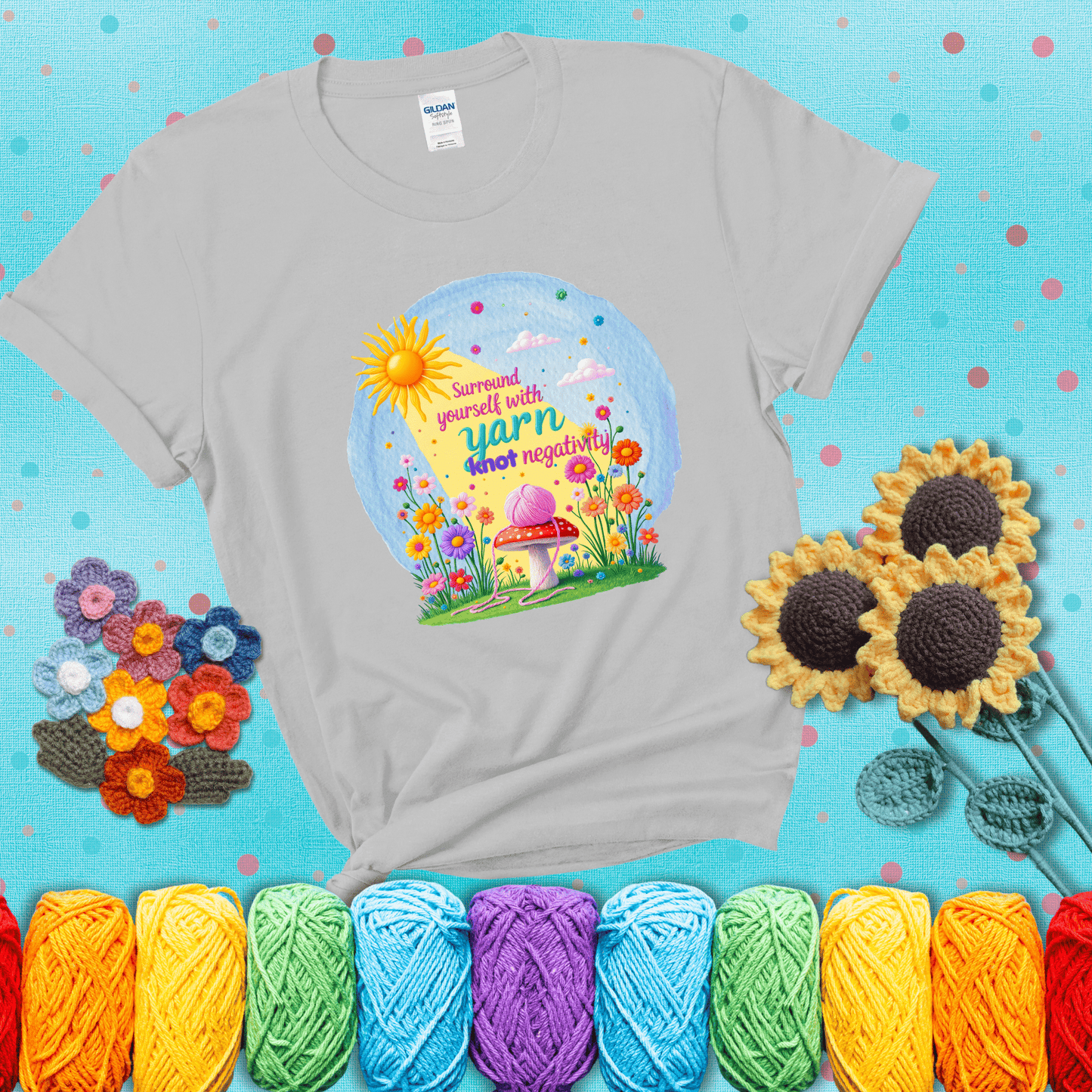 A happy spring garden scene with a sunbeam, flowers, and a pink yarn ball on a mushroom, with the quote "Surround yourself with yarn, knot negativity," showing a light grey tee with a teal sunflower background