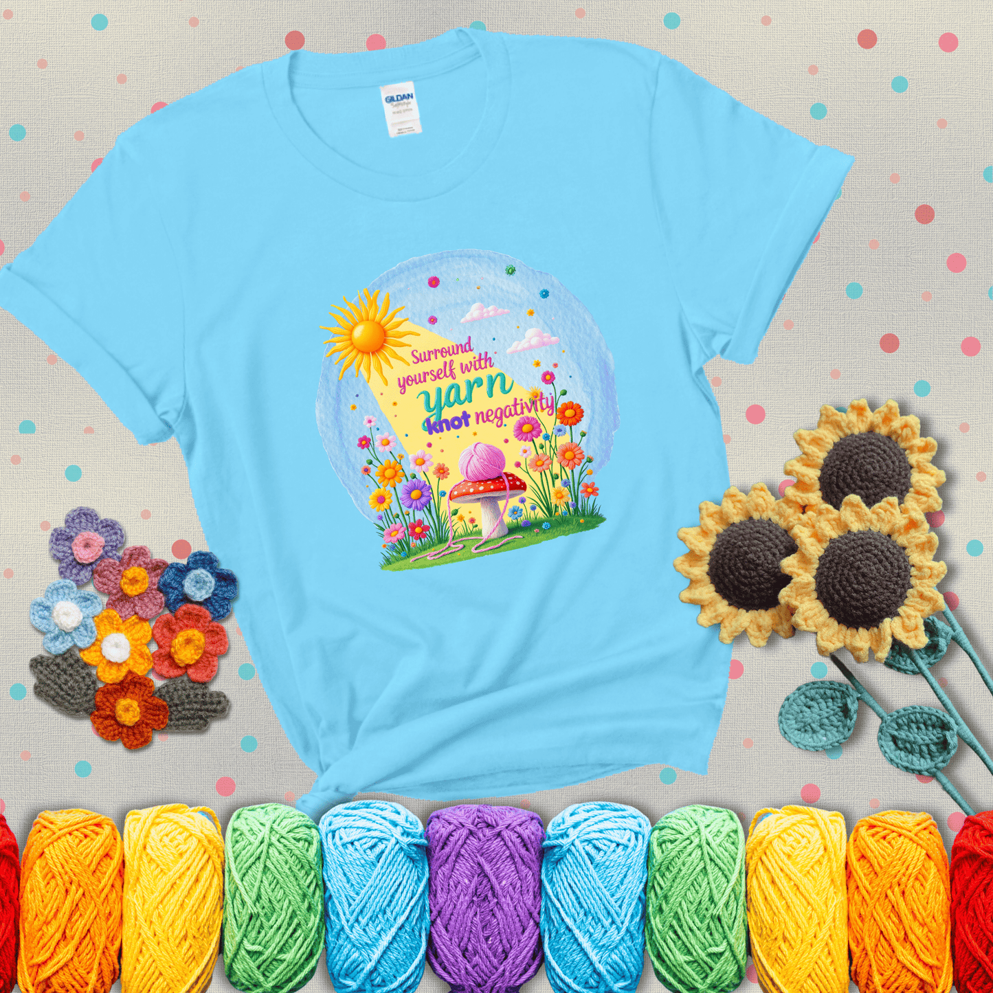 A happy spring garden scene with a sunbeam, flowers, and a pink yarn ball on a mushroom, with the quote "Surround yourself with yarn, knot negativity," showing a light blue tee with grey sunflower background