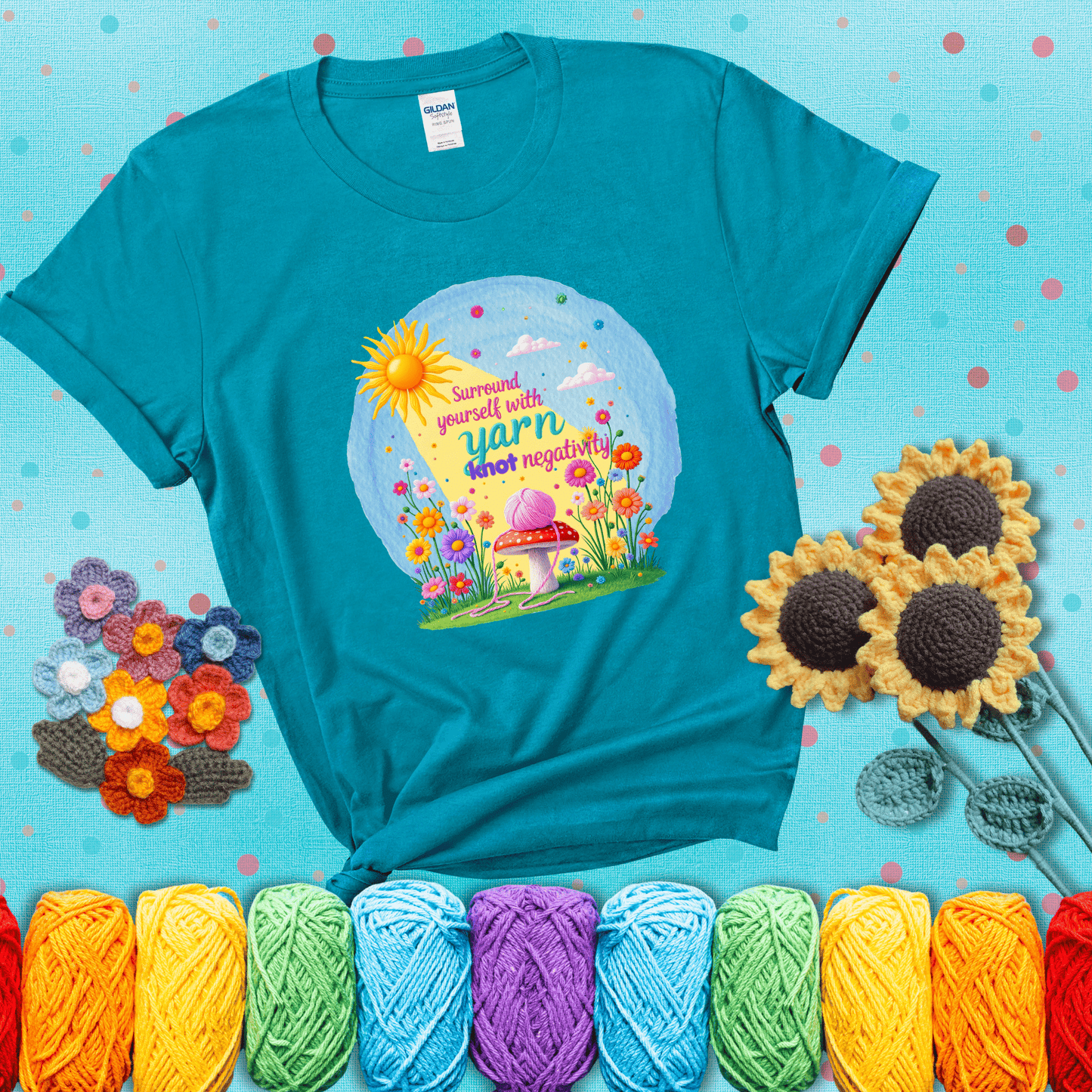 A happy spring garden scene with a sunbeam, flowers, and a pink yarn ball on a mushroom, with the quote "Surround yourself with yarn, knot negativity," showing a blue-green tee with a teal sunflower background