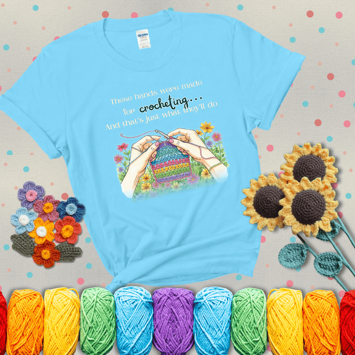 Illustration of two hands crocheting a rainbow swatch, surrounded by vibrant flowers with the phrase “These hands were made for crocheting… and that’s just what they’ll do,” showing a light blue tee with grey sunflower background