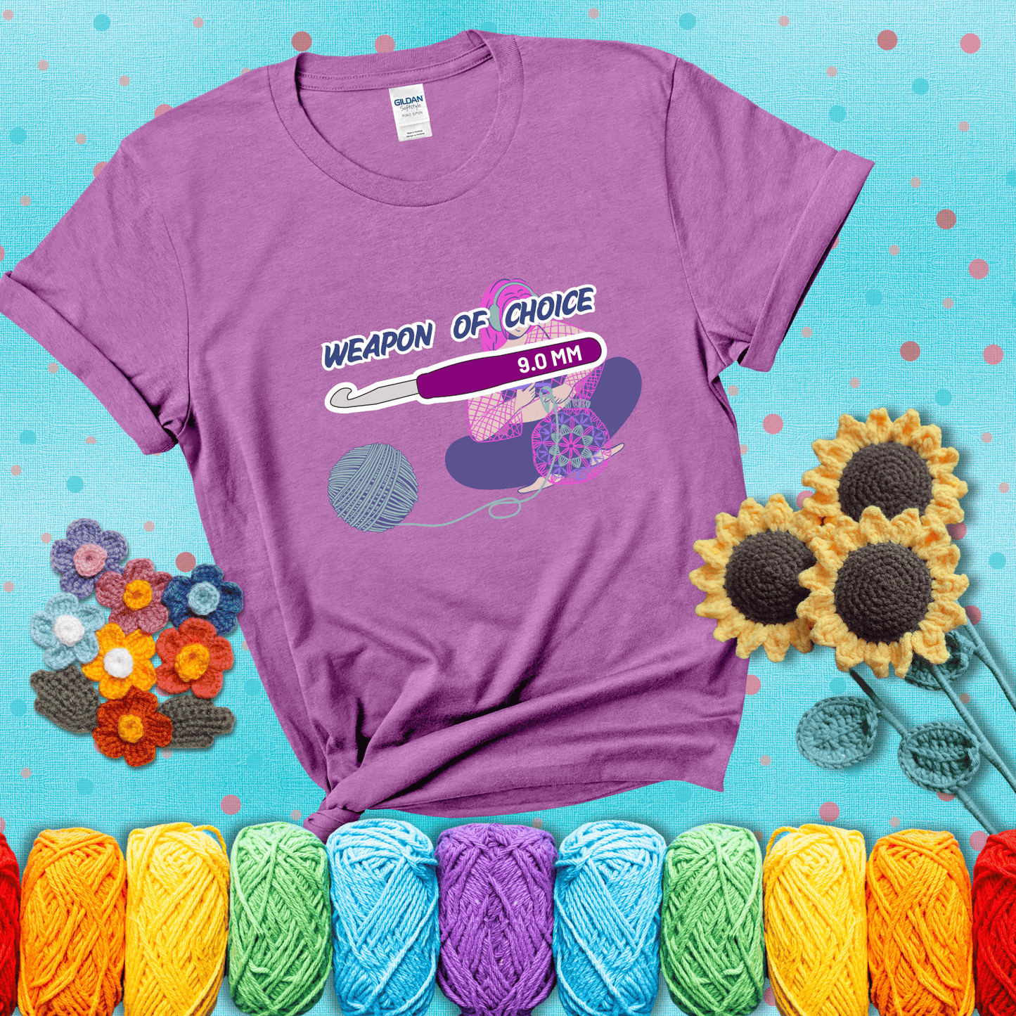 Illustration of a crocheter sitting cross-legged with a 9.0 mm crochet hook above her, labeled “Weapon of Choice.” She’s crocheting a colorful mandala-style project from a large yarn ball, showing a light purple tee with a teal sunflower background