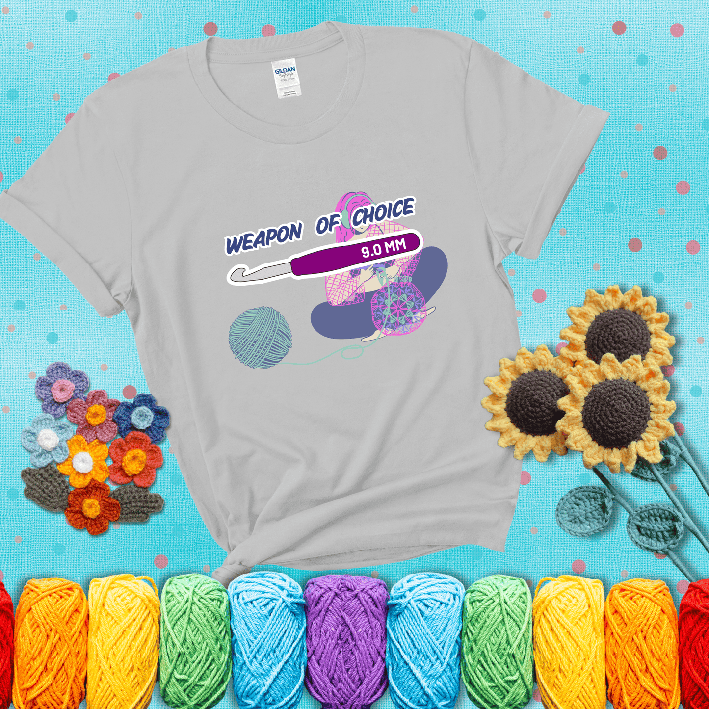 Illustration of a crocheter sitting cross-legged with a 9.0 mm crochet hook above her, labeled “Weapon of Choice.” She’s crocheting a colorful mandala-style project from a large yarn ball, showing a light grey tee with a teal sunflower background