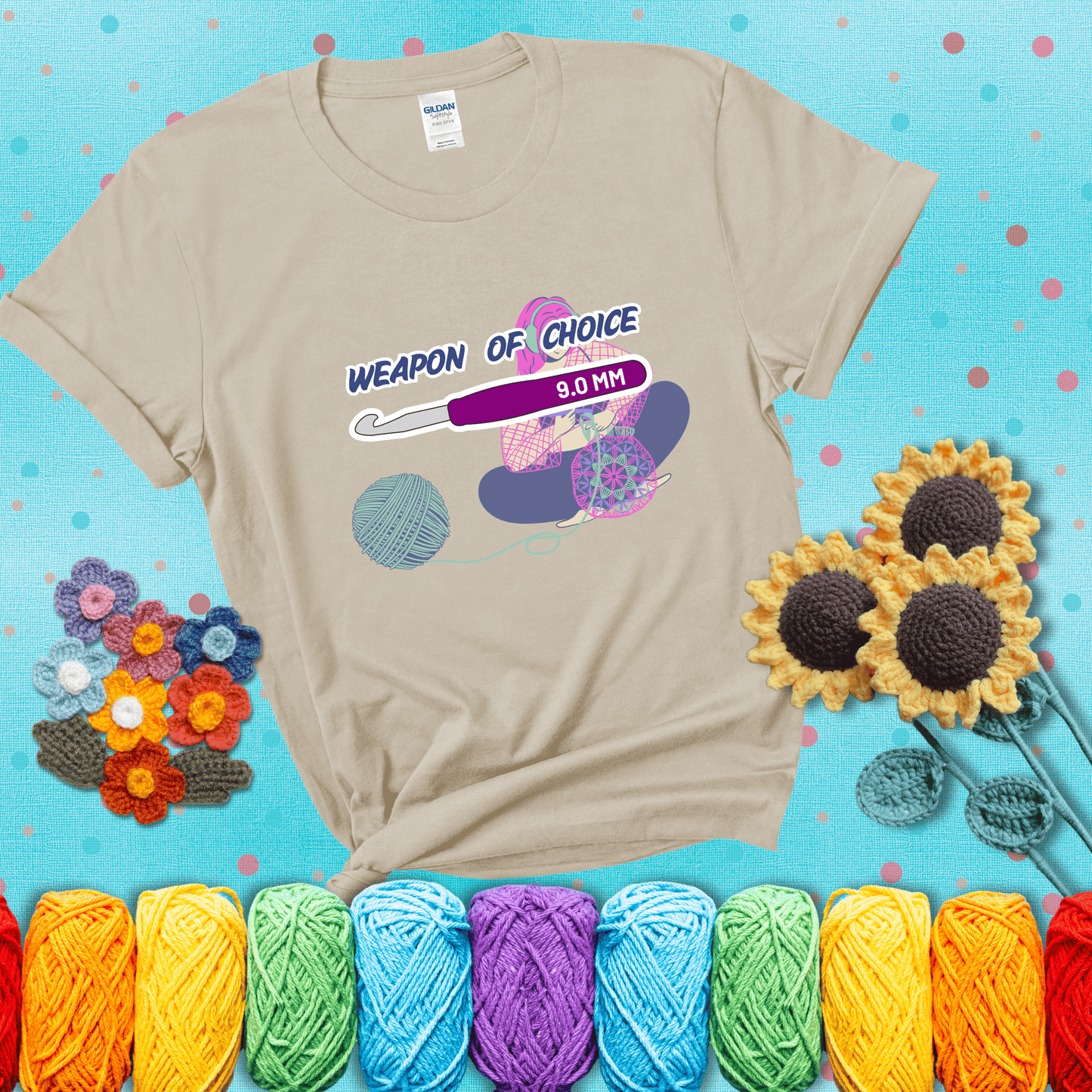Illustration of a crocheter sitting cross-legged with a 9.0 mm crochet hook above her, labeled “Weapon of Choice.” She’s crocheting a colorful mandala-style project from a large yarn ball, showing a beige tee with a teal sunflower background