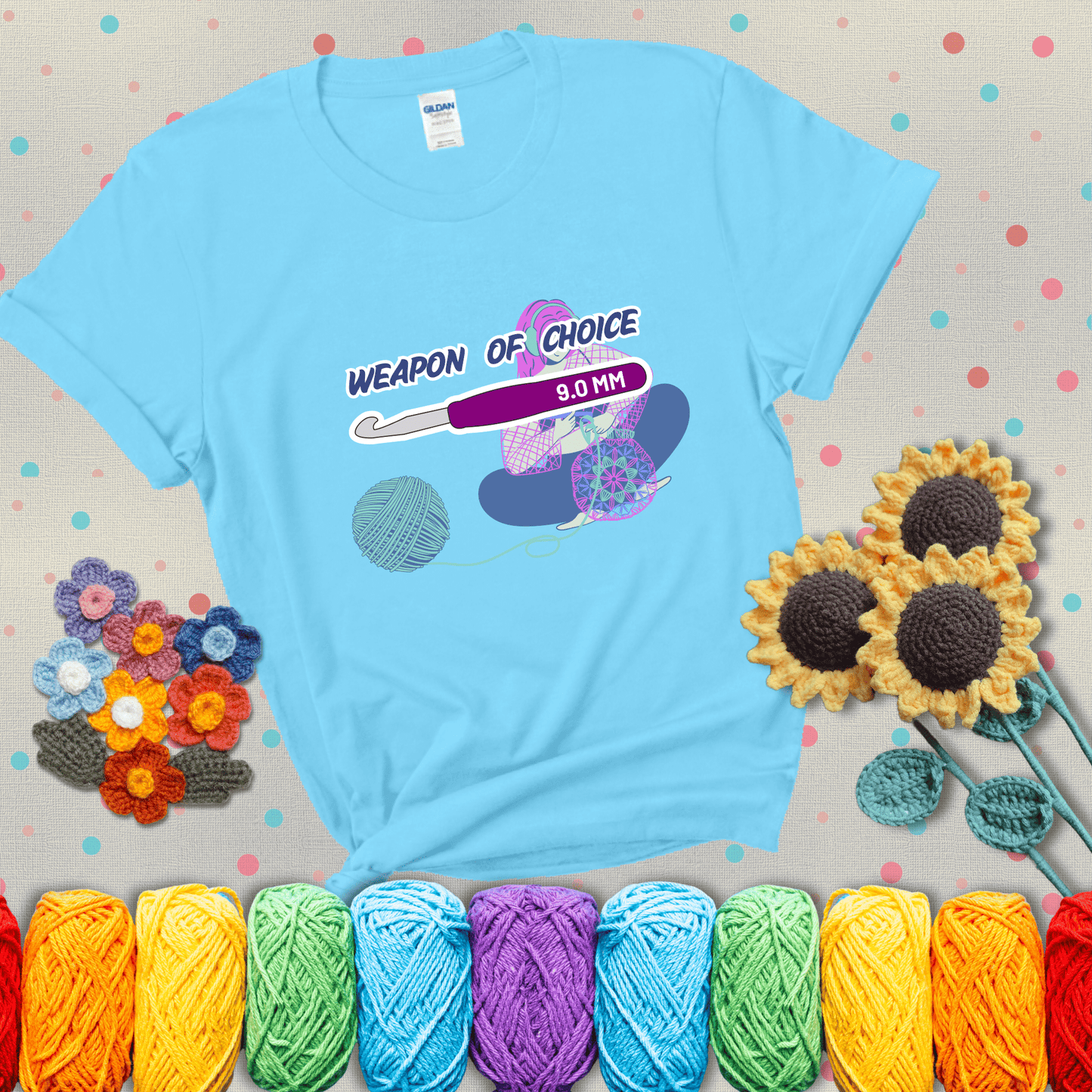 Illustration of a crocheter sitting cross-legged with a 9.0 mm crochet hook above her, labeled “Weapon of Choice.” She’s crocheting a colorful mandala-style project from a large yarn ball, showing a light blue tee with grey sunflower background