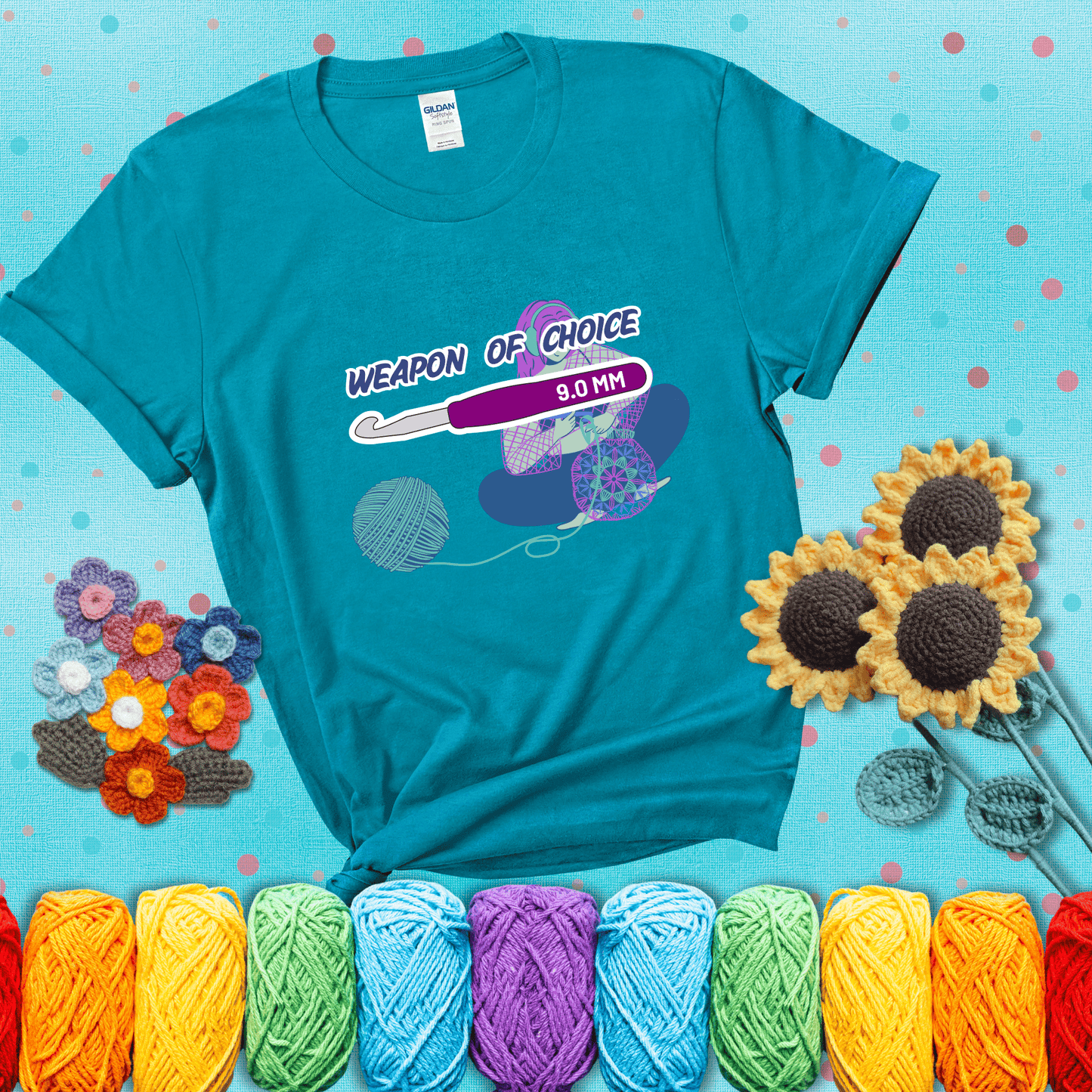 Illustration of a crocheter sitting cross-legged with a 9.0 mm crochet hook above her, labeled “Weapon of Choice.” She’s crocheting a colorful mandala-style project from a large yarn ball, showing a blue-green tee with a teal sunflower background