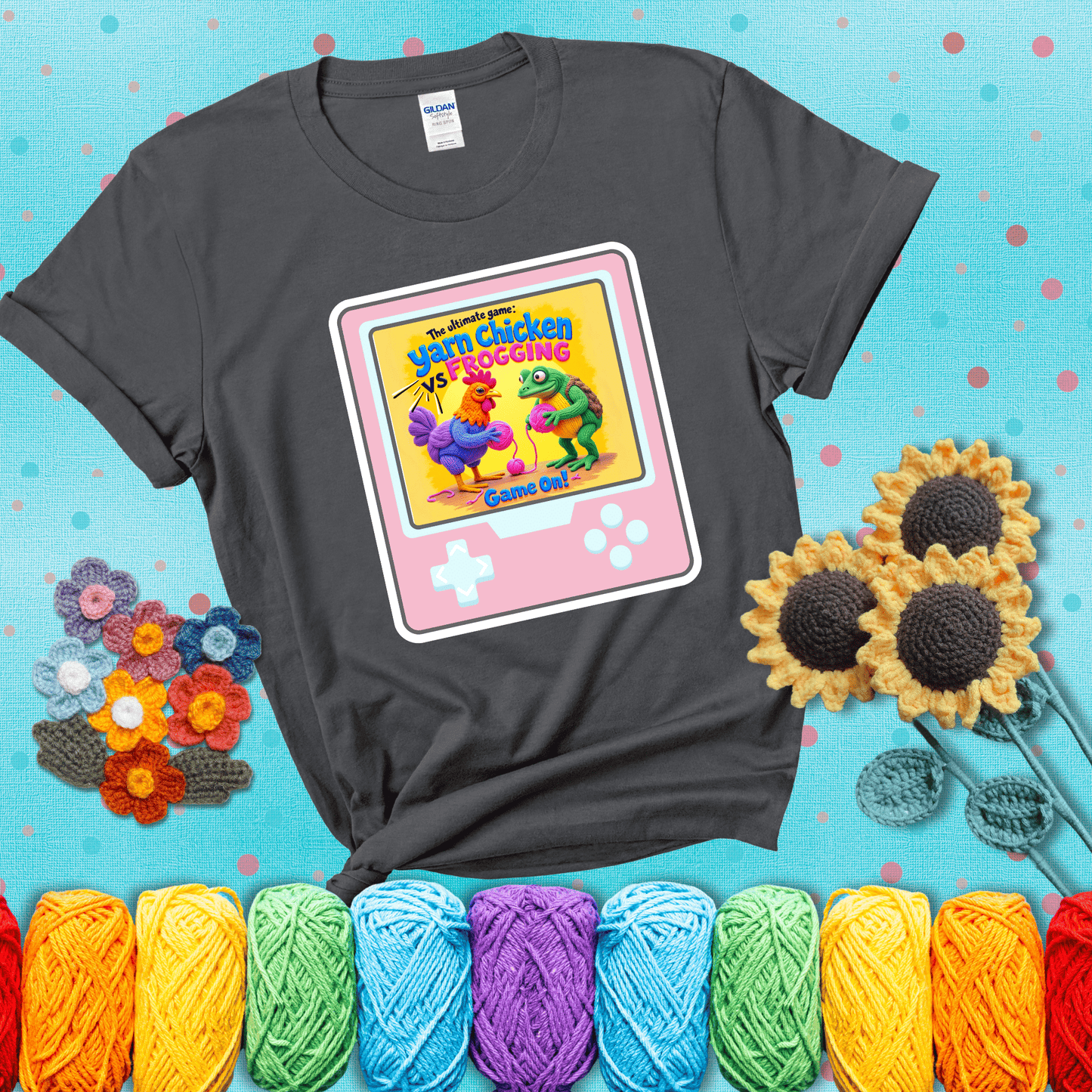 A colorful chicken and frog holding pink yarn face off inside a retro handheld game screen with the title “The ultimate game: Yarn Chicken vs. Frogging – Game On!” showing a dark grey tee with a teal sunflower background