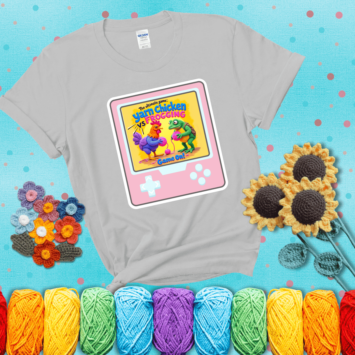 A colorful chicken and frog holding pink yarn face off inside a retro handheld game screen with the title “The ultimate game: Yarn Chicken vs. Frogging – Game On!” showing a light grey tee with a teal sunflower background