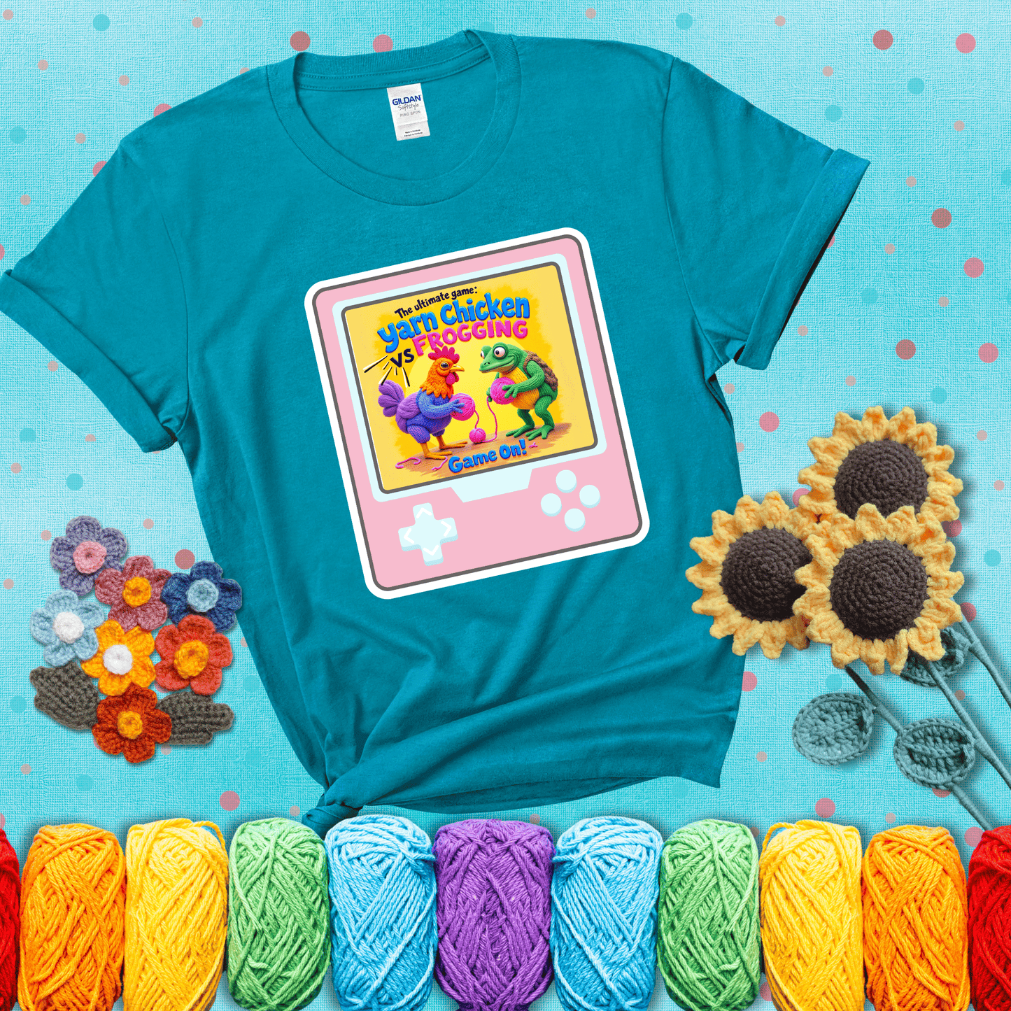 A colorful chicken and frog holding pink yarn face off inside a retro handheld game screen with the title “The ultimate game: Yarn Chicken vs. Frogging – Game On!” showing a blue-green tee with a teal sunflower background