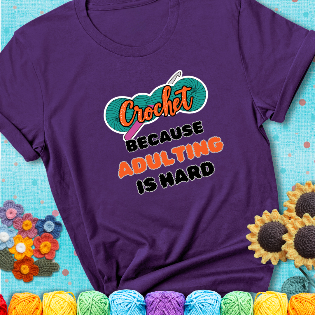 Purple t-shirt featuring a bold design with the phrase “Crochet Because Adulting Is Hard” featuring a green skein of yarn with a pink crochet hook. The word “Crochet” is in a script font, while “Adulting is hard” is in chunky textured letters.