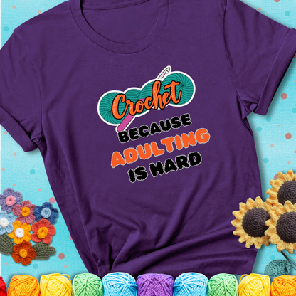 Purple t-shirt featuring a bold design with the phrase “Crochet Because Adulting Is Hard” featuring a green skein of yarn with a pink crochet hook. The word “Crochet” is in a script font, while “Adulting is hard” is in chunky textured letters.