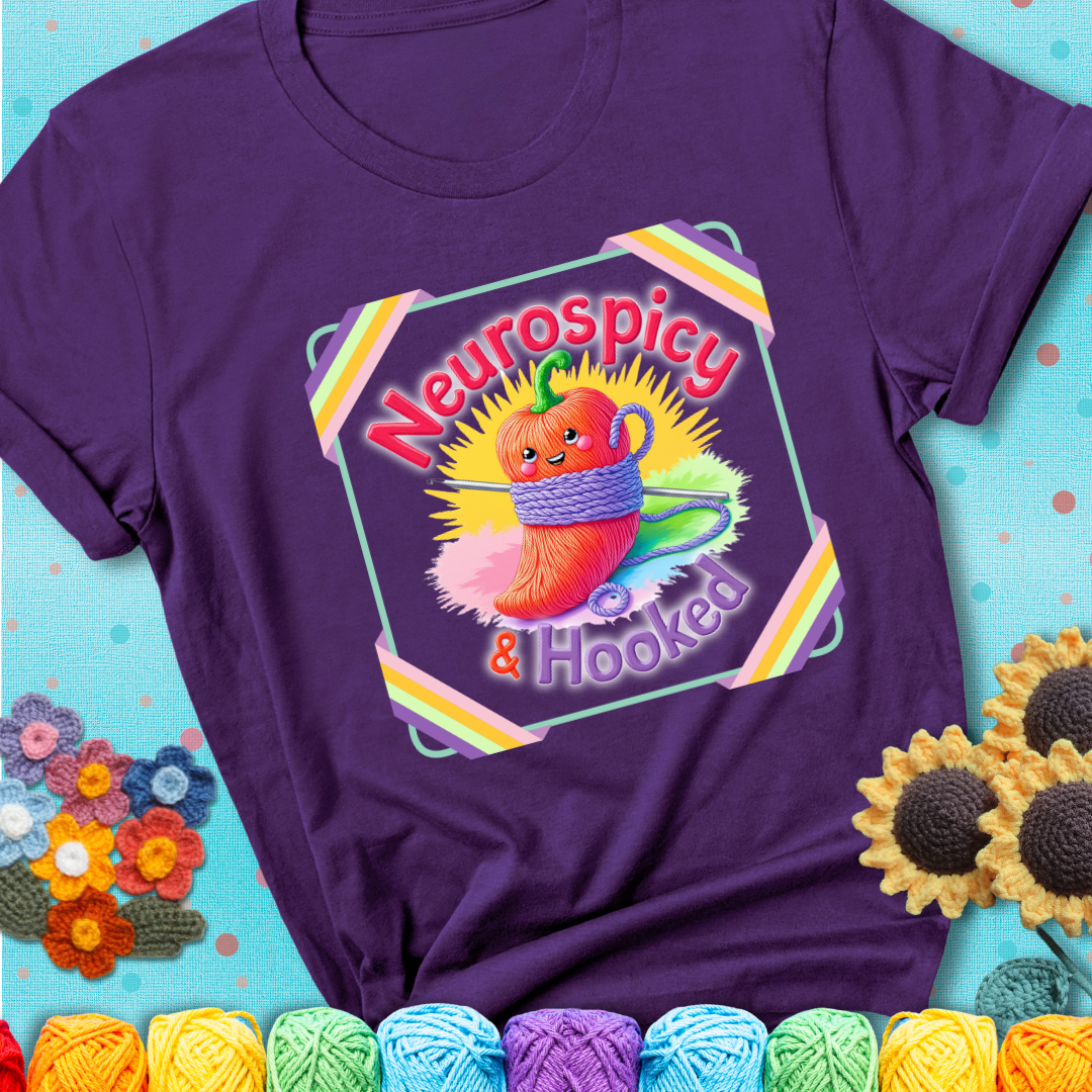 Purple t-shirt featuring a smiling red chili pepper wrapped in yarn with a crochet hook, text says “Neurospicy & Hooked.”