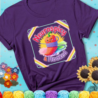 Purple t-shirt featuring a smiling red chili pepper wrapped in yarn with a crochet hook, text says “Neurospicy & Hooked.”