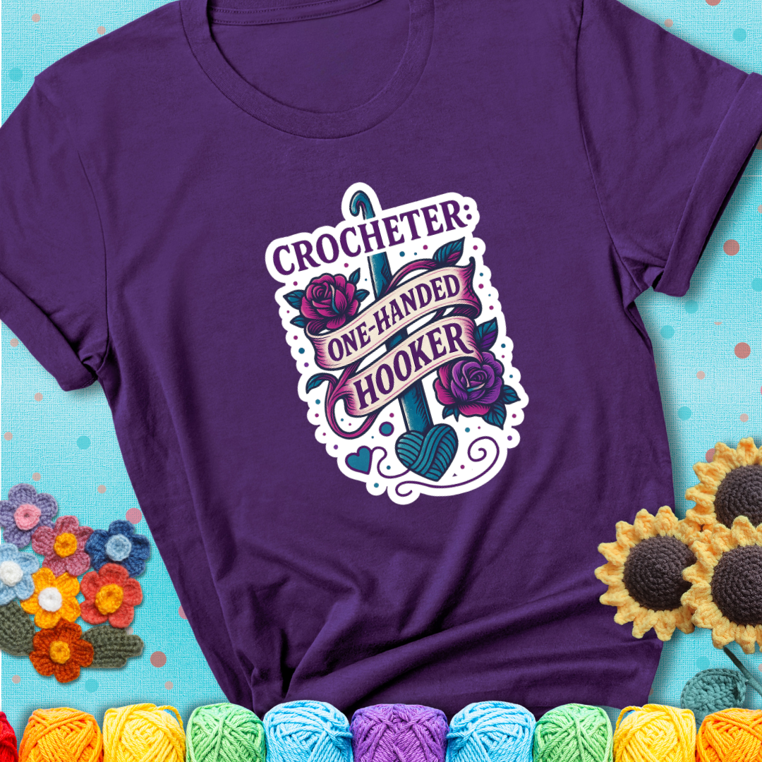 Purple t-shirt with a tattoo-style crochet design featuring a crochet hook, roses, and the phrase “Crocheter: One-Handed Hooker.”