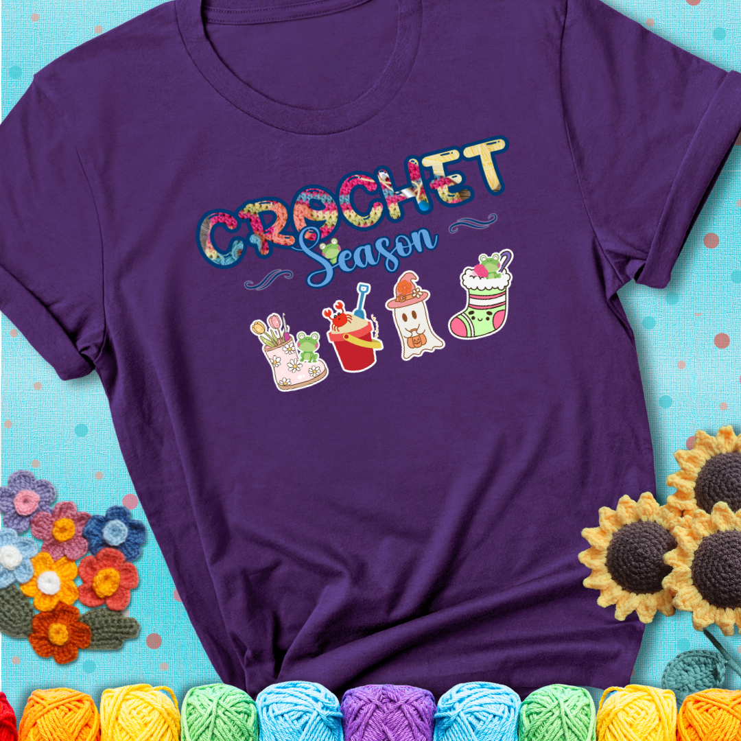 Purple t-shirt shows the words “Crochet Season” with cute seasonal icons below: a spring boot with hooks, summer bucket with crab, fall ghost with pumpkin hat, winter stocking, and tiny frogs.