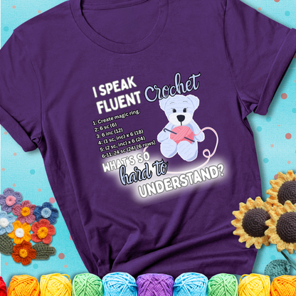 A cute crochet bear holding a yarn ball, surrounded by a short crochet pattern. The phrase reads “I Speak Fluent Crochet – What’s so hard to understand?” on a purple t-shirt.
