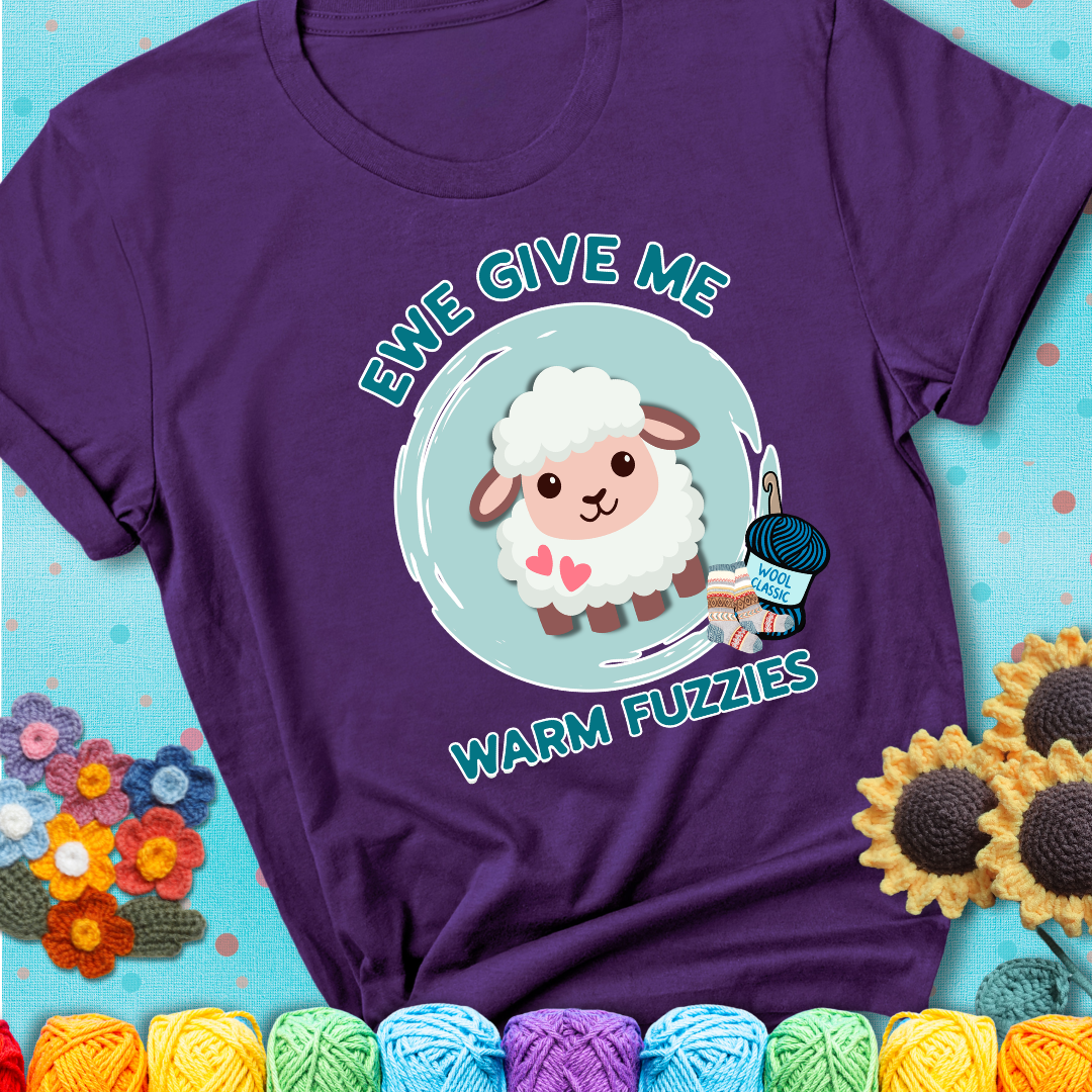 Purple t-shirt showing a cartoon sheep with pink hearts, wool socks, and yarn labeled “Wool Classic.” Text reads “Ewe Give Me Warm Fuzzies.”