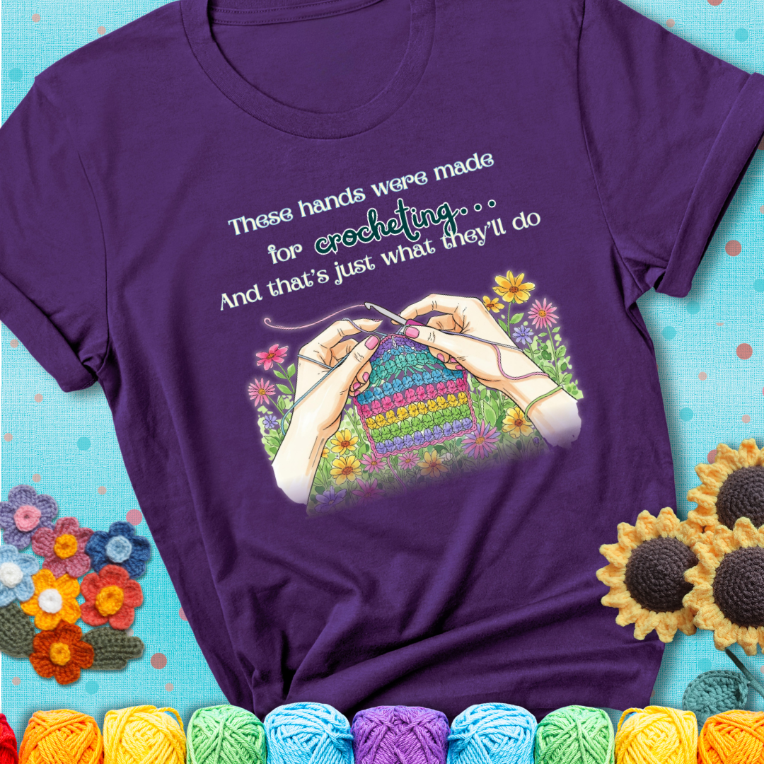 Purple t-shirt with an illustration of two hands crocheting a rainbow swatch, surrounded by colorful flowers and the phrase “These hands were made for crocheting… and that’s just what they’ll do.”