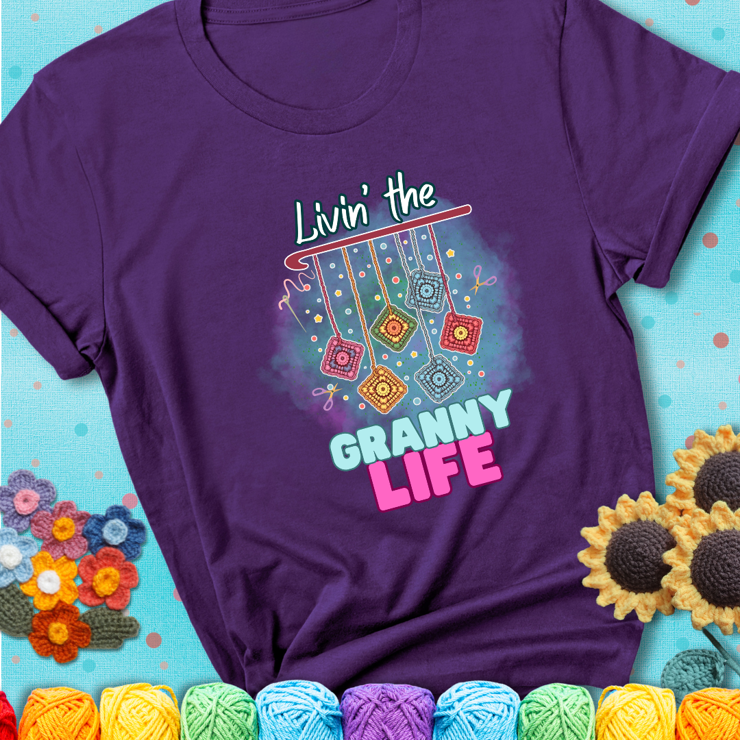 Purple t-shirt shows colorful crochet granny squares hanging from a hook with text “Livin’ the Granny Life” in bold pink and blue font; a fun nod to classic crochet.
