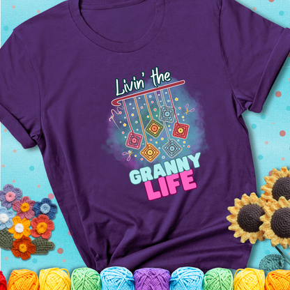 Purple t-shirt shows colorful crochet granny squares hanging from a hook with text “Livin’ the Granny Life” in bold pink and blue font; a fun nod to classic crochet.