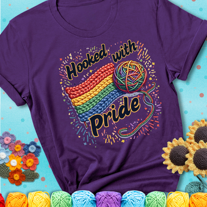 A rainbow-striped crochet swatch paired with a multicolor yarn ball and bold “Hooked With Pride” lettering. Confetti surrounds the vibrant scene showing a purple tee with a teal sunflower background