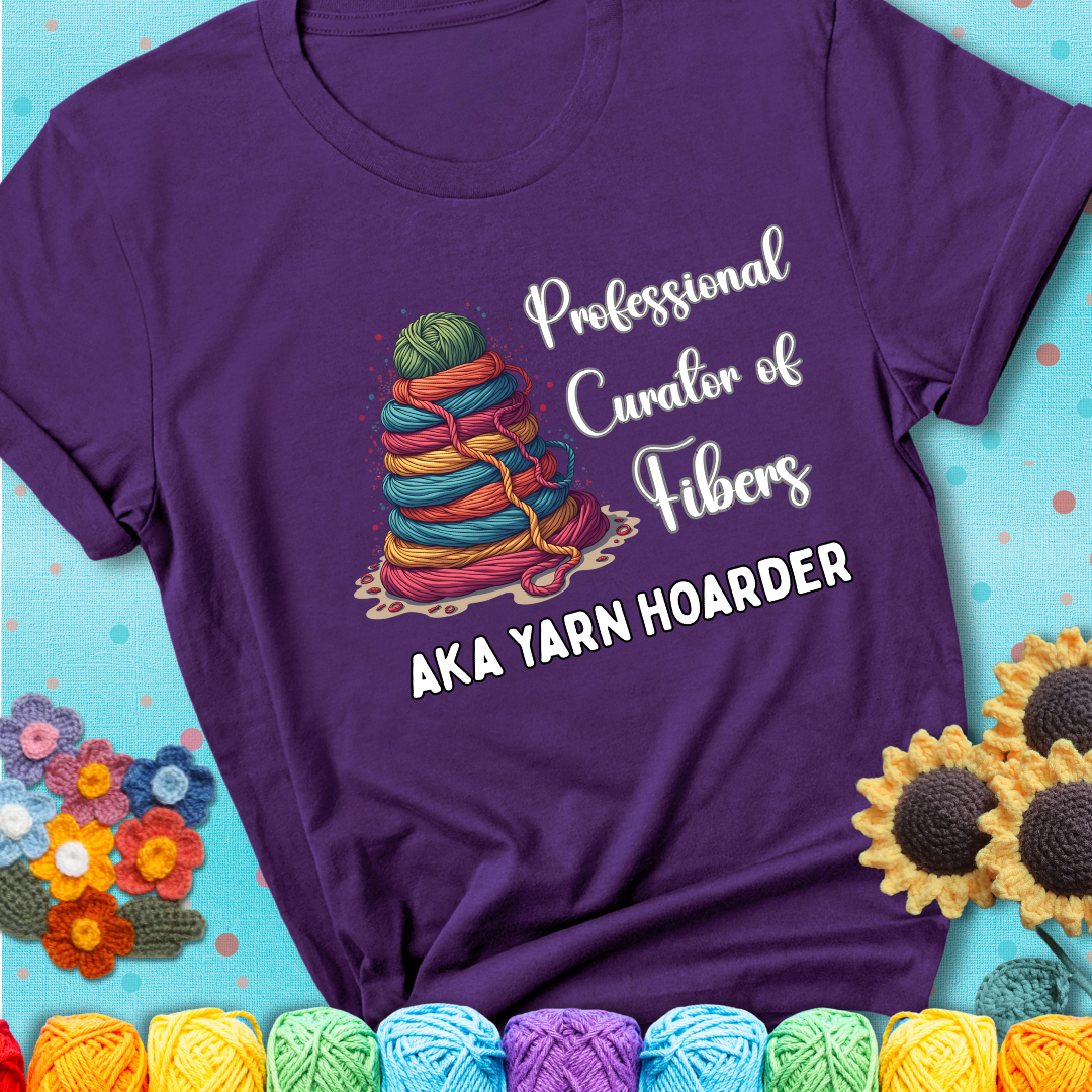 A purple t-shirt showing a pyramid of rainbow yarn topped with green ball; text reads “Professional Curator of Fibers AKA Yarn Hoarder”, a playful shirt for serious stashers.