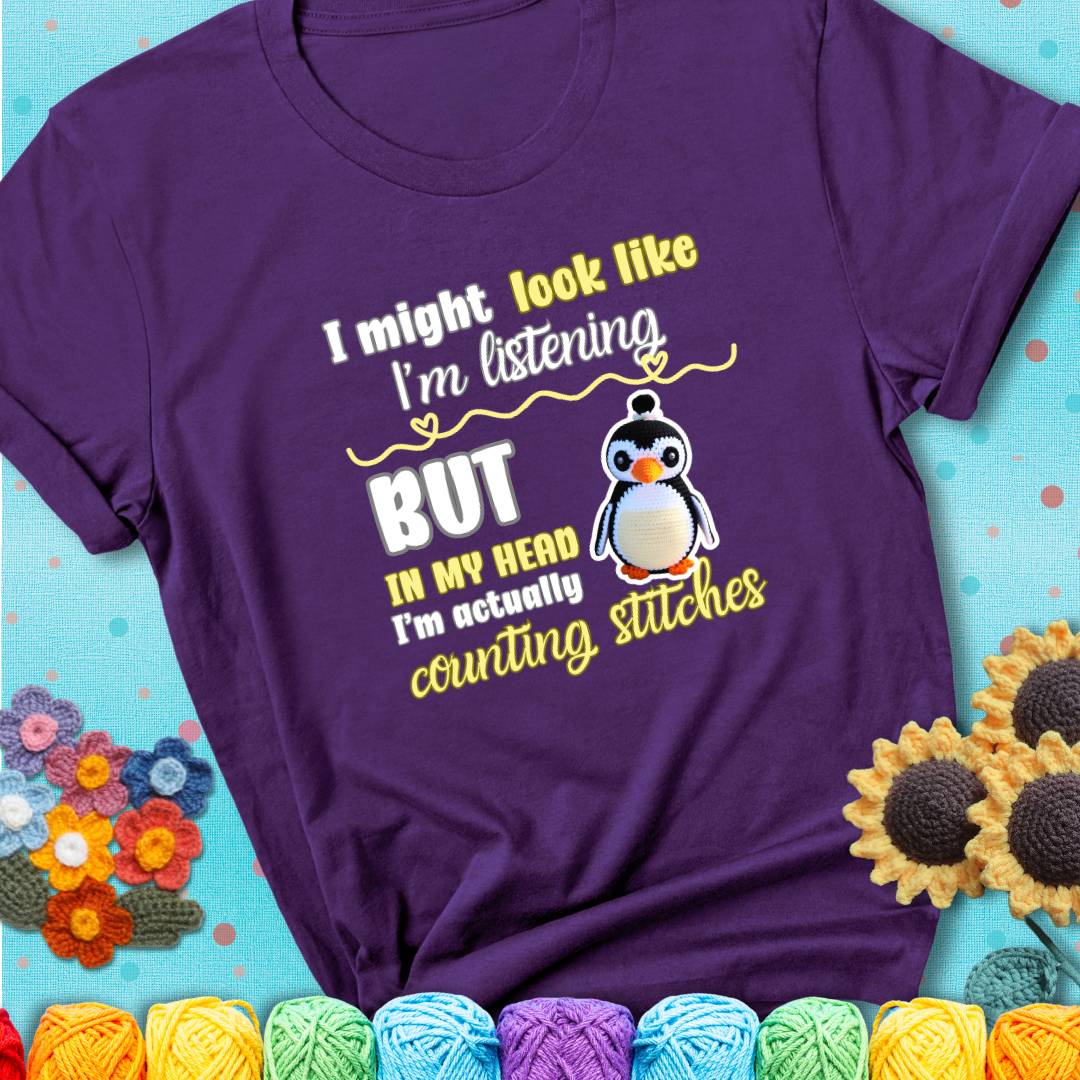 Purple t-shirt shows a crocheted penguin next to the text “I might look like I’m listening but in my head I’m actually counting stitches.”