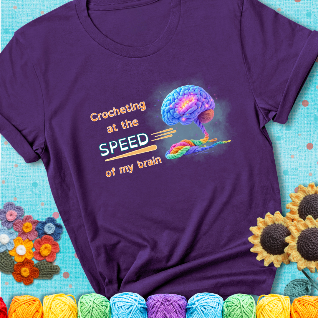 A glowing rainbow brain unravels into colorful yarn with the phrase “Crocheting at the speed of my brain” on a purple t-shirt. 