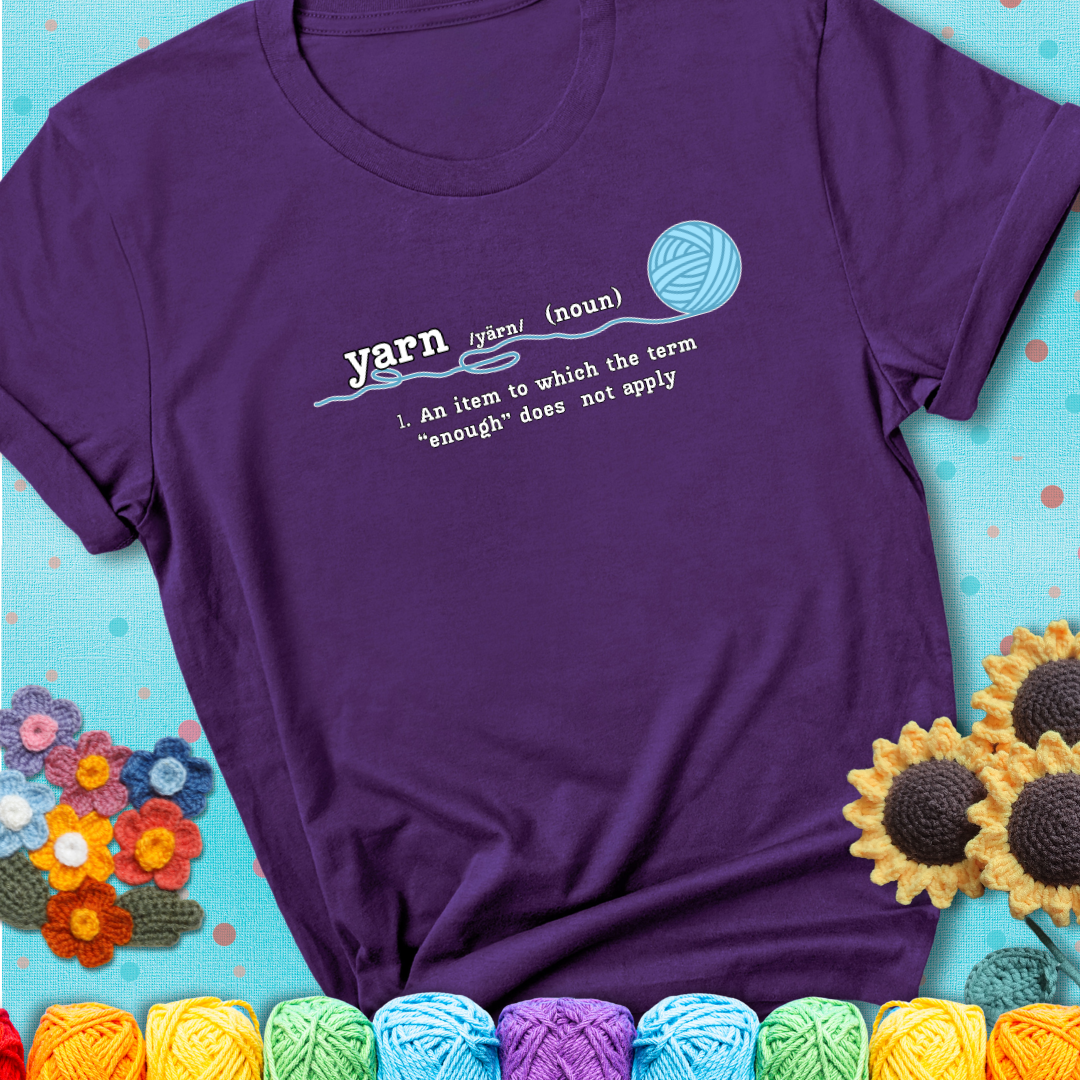 Purple blue t-shirt with a definition-style yarn graphic reading “An item to which the word ‘enough’ does not apply,” plus a blue ball of yarn with a loose strand.