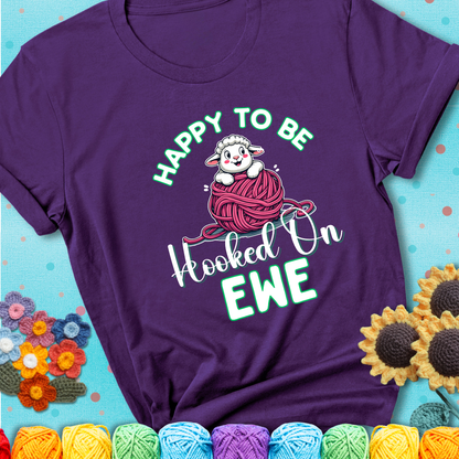 Purple t-shirt with smiling cartoon sheep peeks from a pink yarn ball, surrounded by green text “Happy to Be Hooked on Ewe”, a playful and affectionate sheep pun for crocheters.