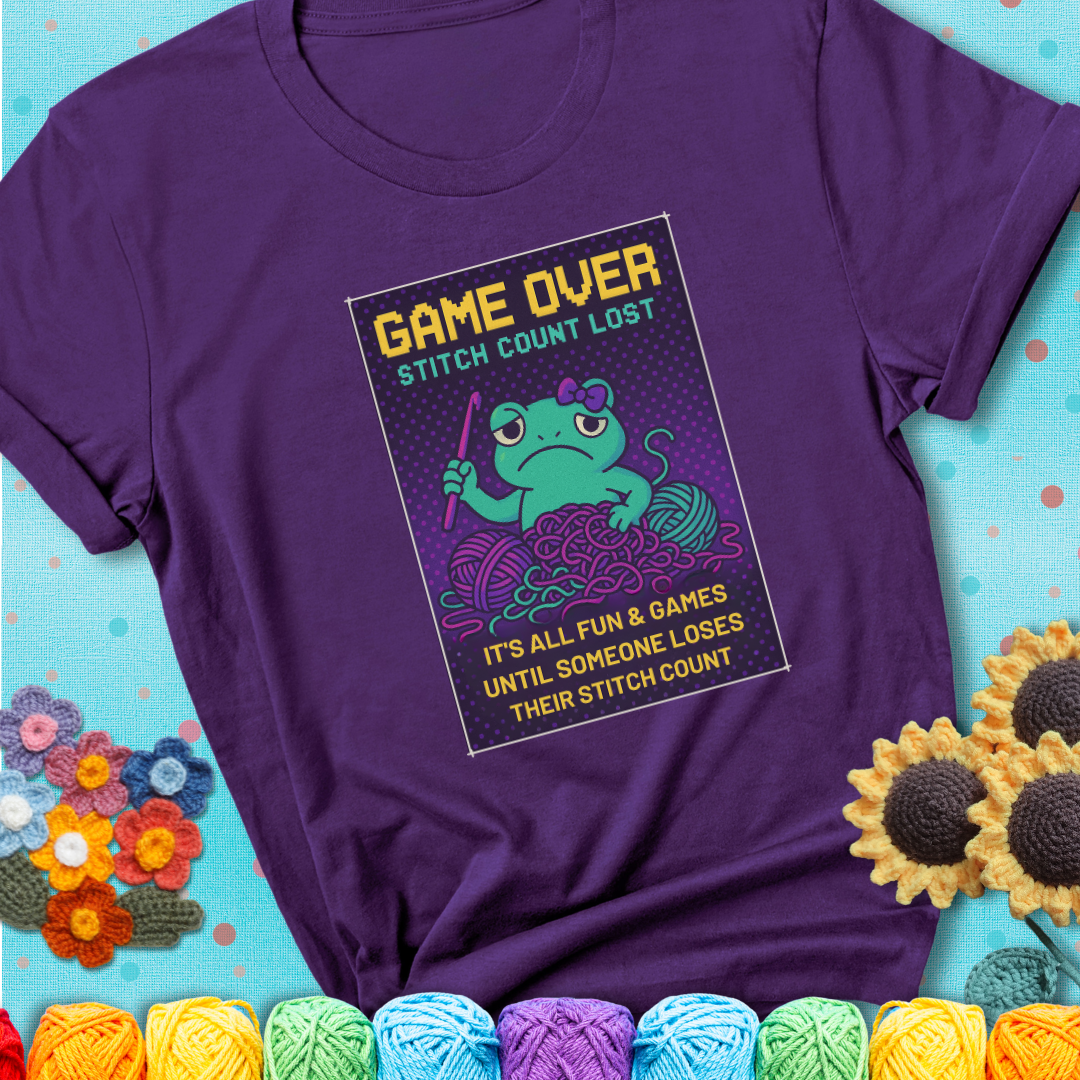 Purple t-shirt shows a pixel-style design of a grumpy yarn frog holding a hook, surrounded by yarn chaos, with the text “Game Over. Stitch Count Lost.”